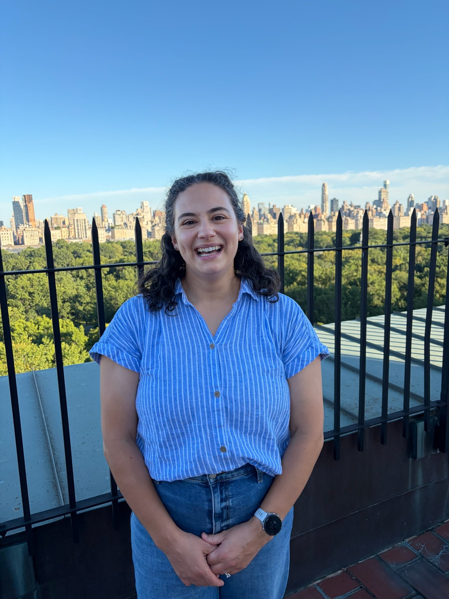 A woman smiling outdoors on a rooftop with the city skyline in the background, wearing a blue striped shirt and a watch.