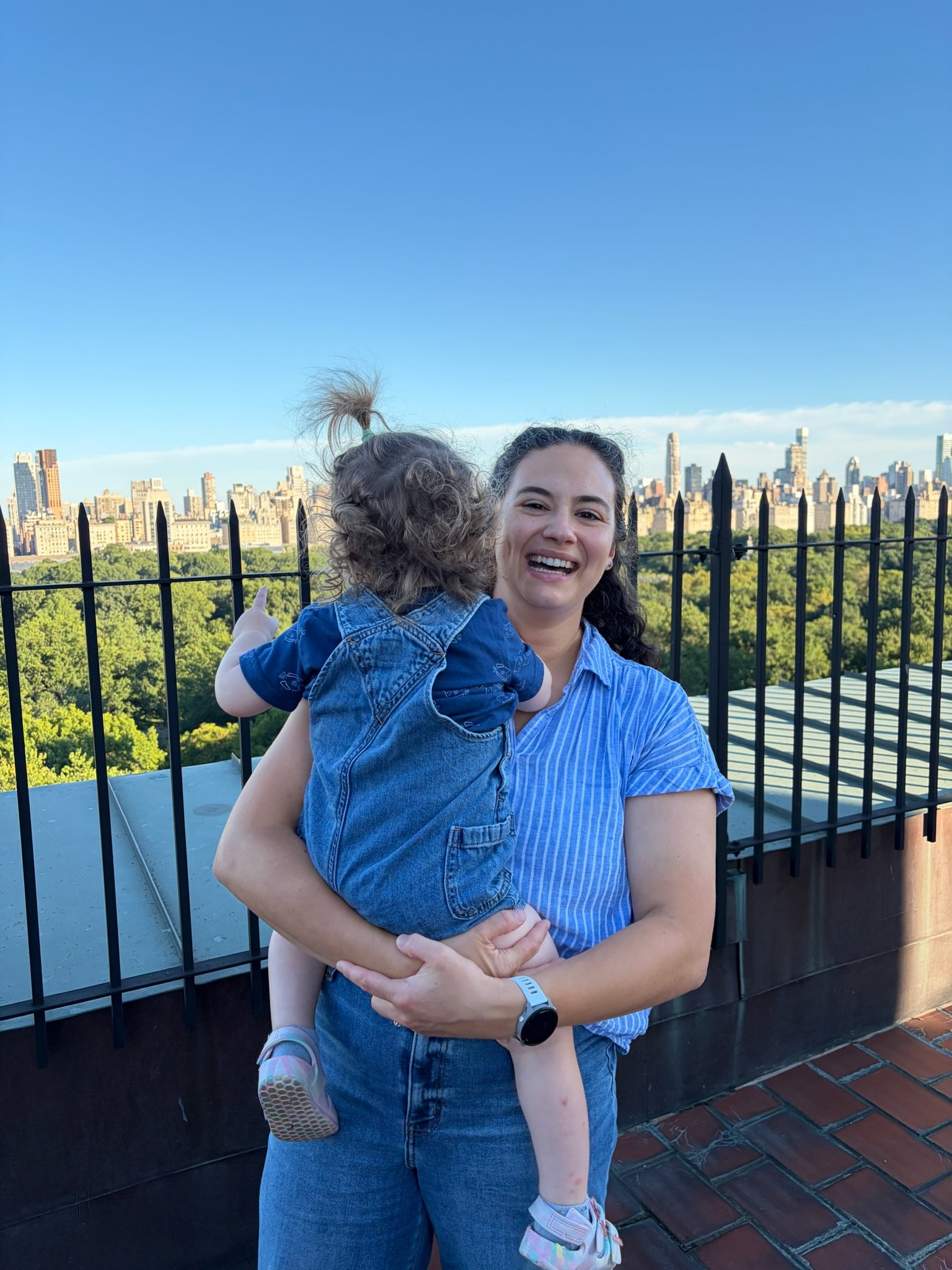 A woman holding a young girl in her arms, standing on a rooftop with a city skyline and green park in the background. The woman is smiling and wearing a blue striped shirt; the girl is wearing a denim jacket and shorts.