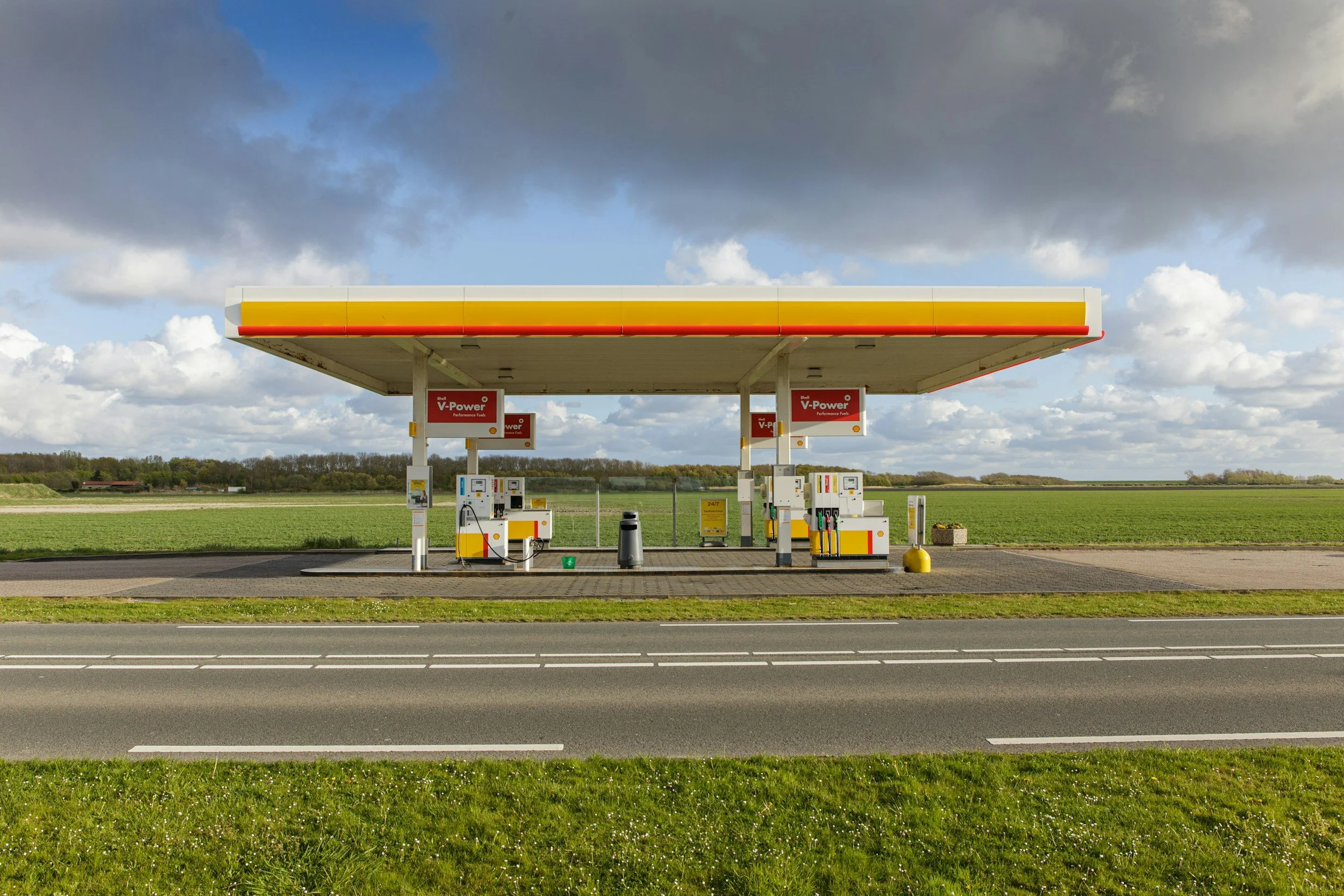 Empty Shell gas station with four fuel pumps under a large yellow and red canopy, set in a rural area with a grassy field and cloudy sky in the background.
