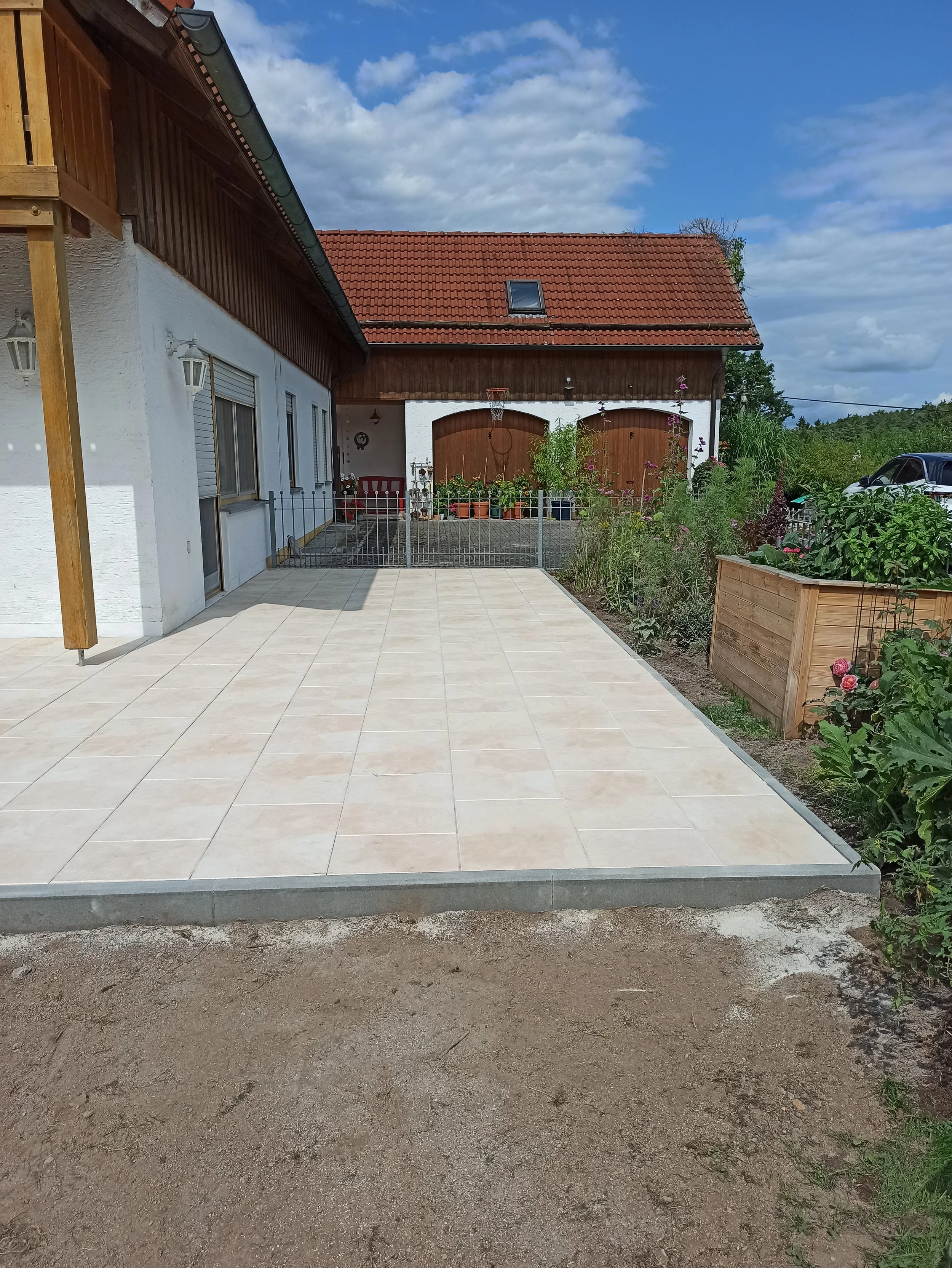 Ein gepflegter Außenbereich eines Hauses mit einer frisch gepflasterten Terrasse, einem Gartenzwerg und einem Blumenkübel mit Pflanzen, im Hintergrund eine Garage mit einer Basketballkörung, an einem windigen Tag bei bewölktem Himmel.