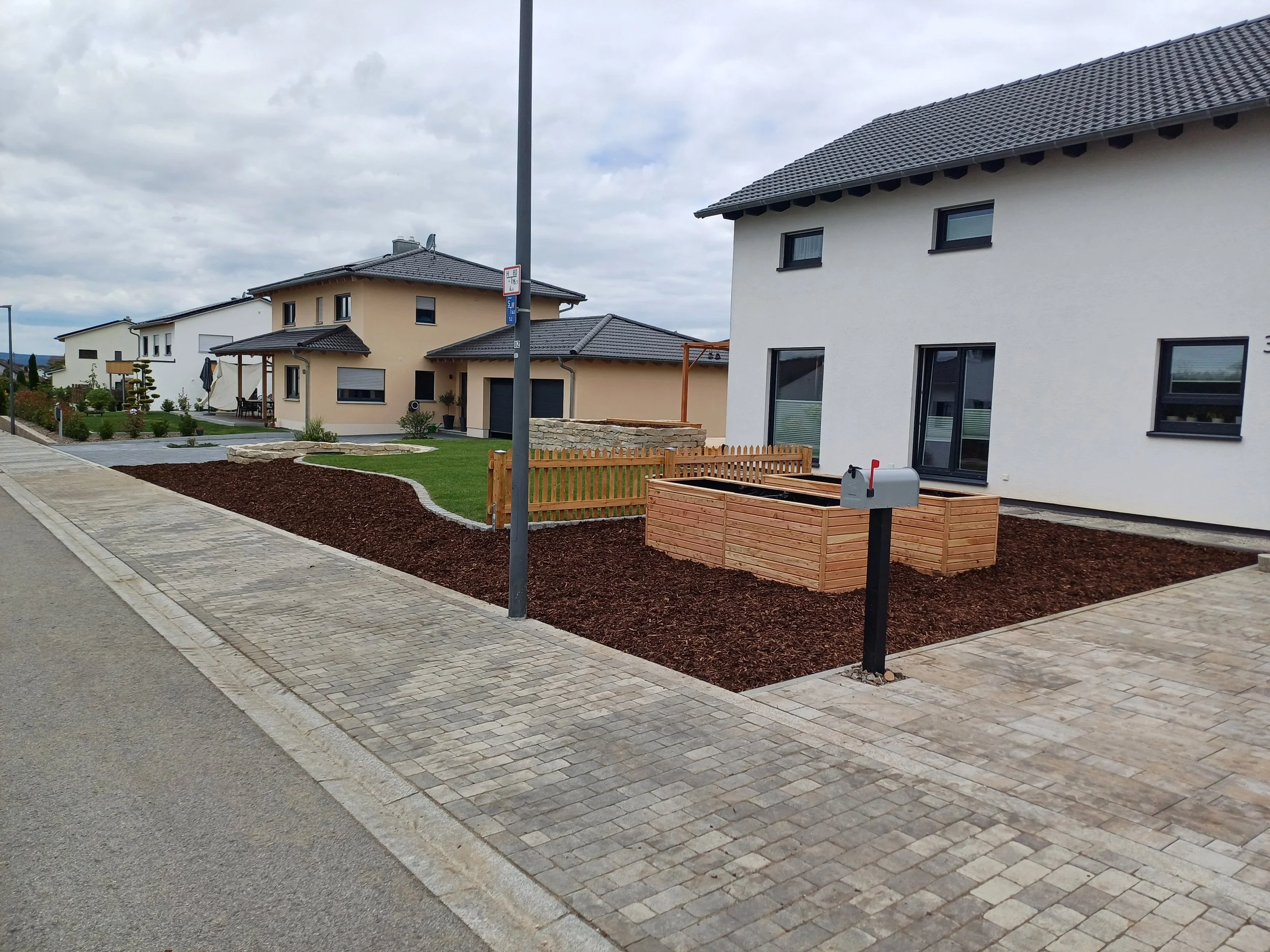 Garten vor einem modernen Haus mit Holzbeeten, kleiner Holzzäunchen, Briefkasten und Parkverbotsschilder, gepflasterter Gehweg und schneebedeckte, bewölkte Himmel.