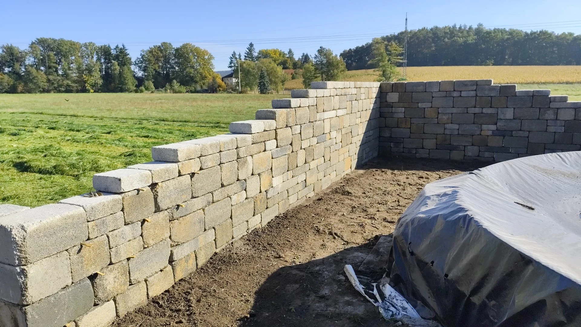Bau einer Steinmauer auf einem Feld mit Bäumen und Hügel im Hintergrund, sonniger Tag.