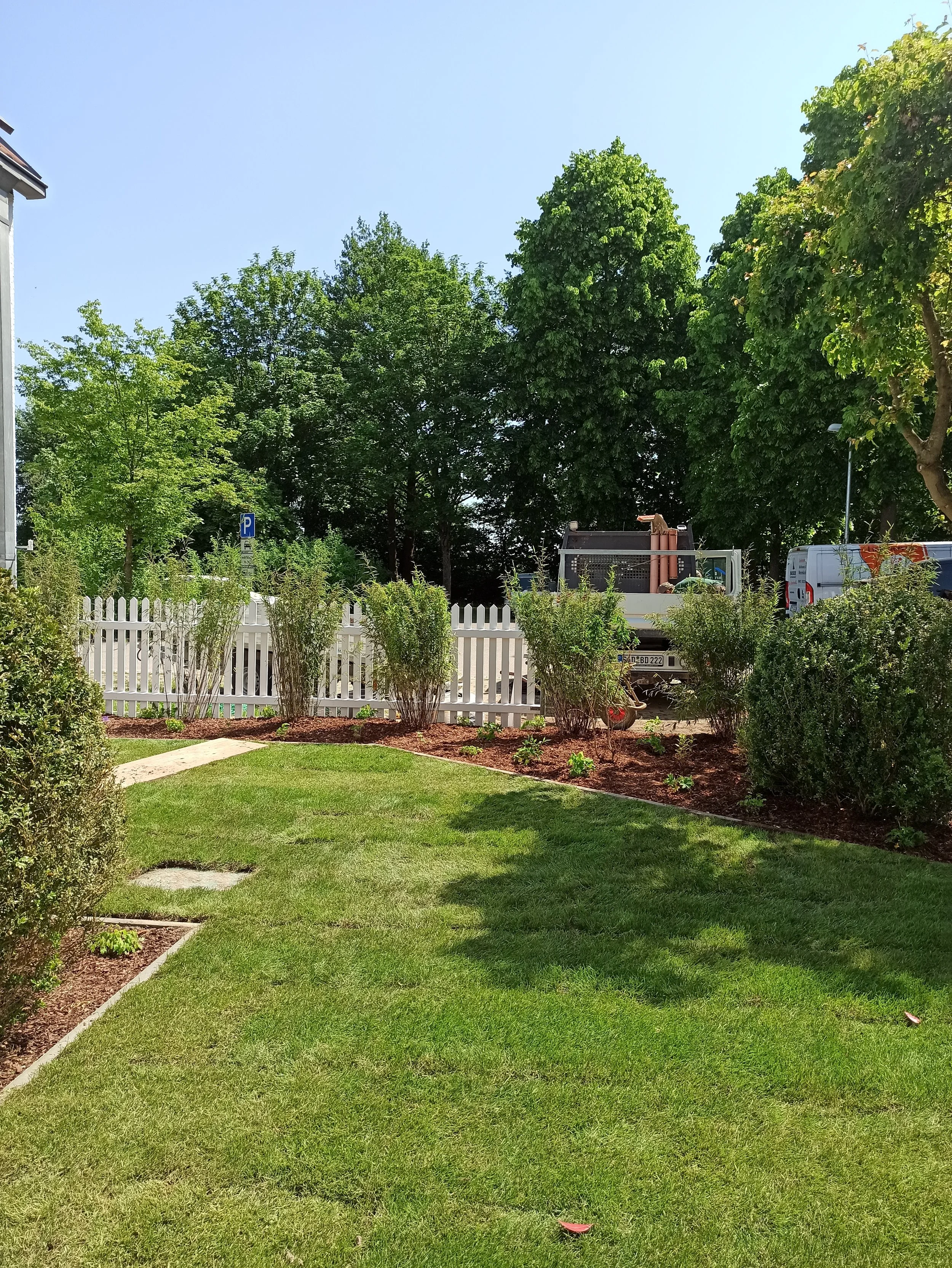 Garten mit grünem Rasen, Büschen, weißen Zaun und Bauwagen im Hintergrund, bei sonnigem Wetter.