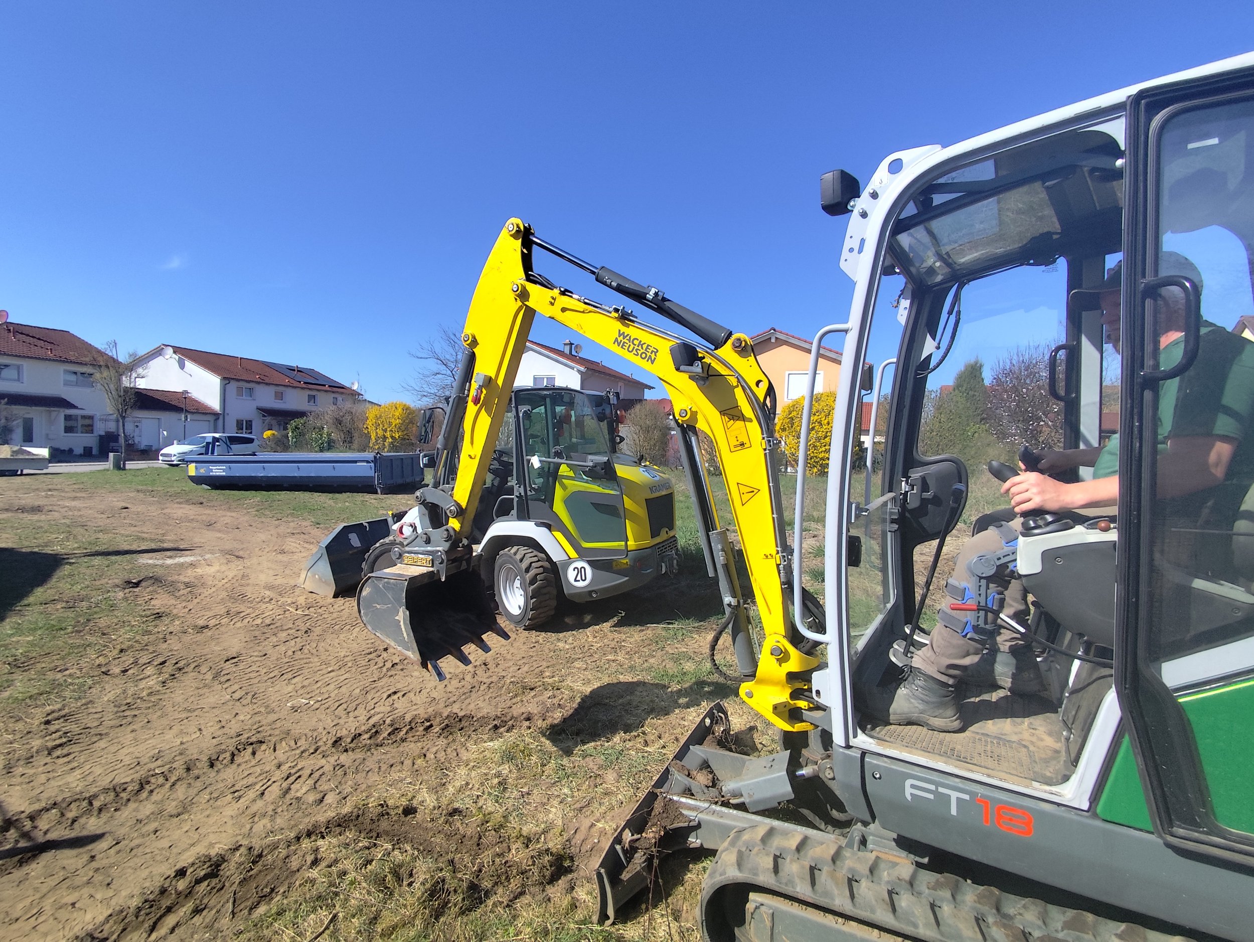 Kleines gelbes Bagger auf einer Baustraße, mit einem Fahrer im geschlossenen Kabinen, beim Arbeiten an einem Bauvorhaben in einem Wohngebiet.