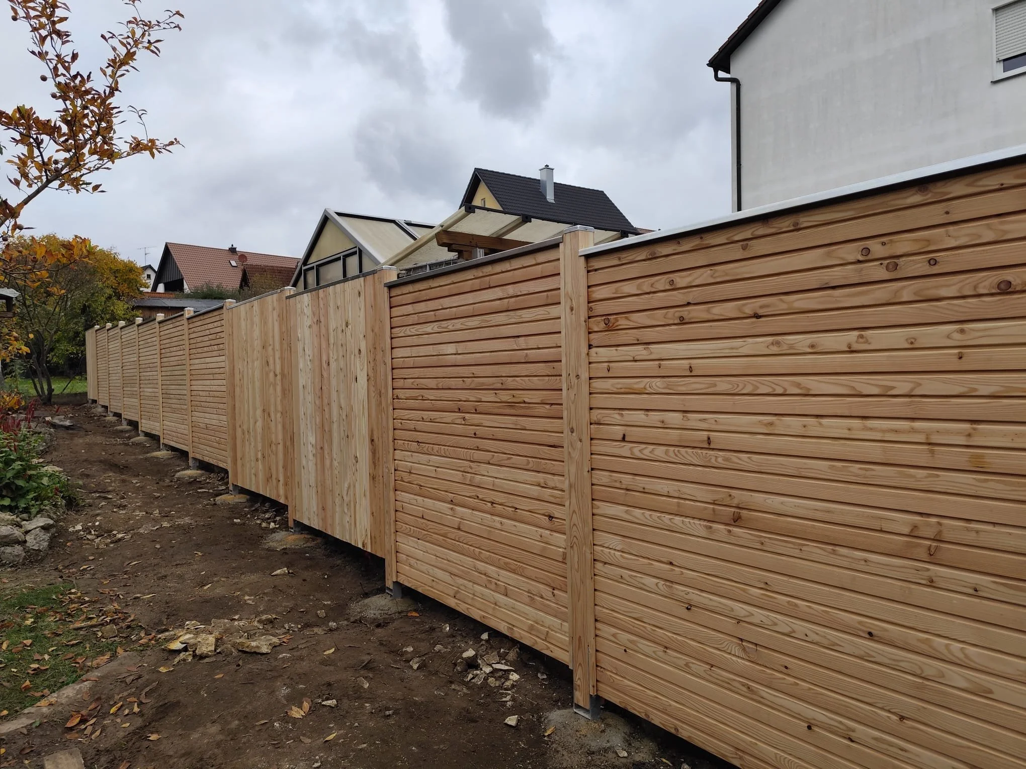Holzlattenzaun in einem Garten, im Hintergrund Häuser bei bewölktem Himmel.