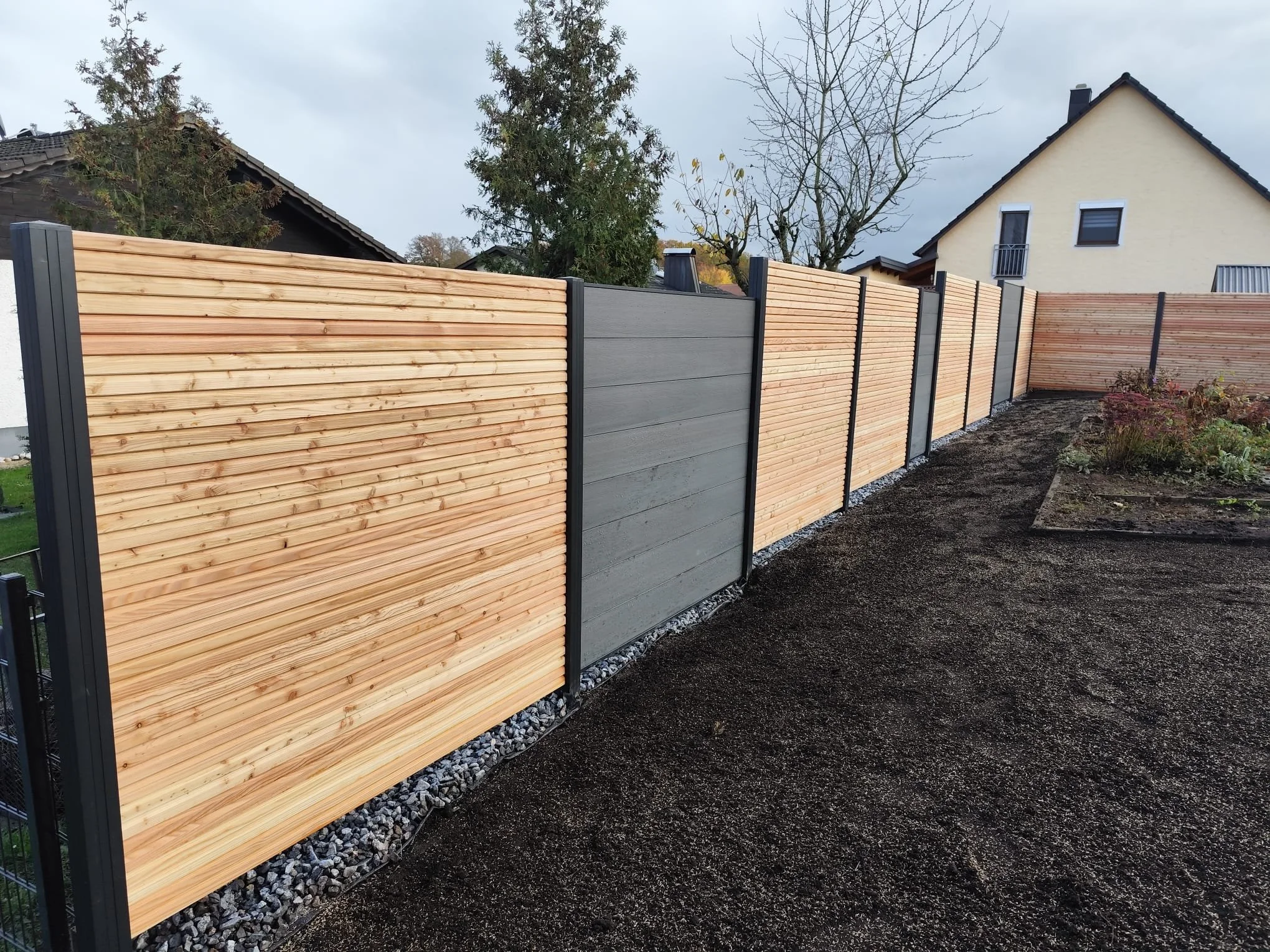 Aufbau eines Zauns mit Holzbrettern und Metallpfosten an einem Garten, im Hintergrund Bäume und Häuser, regnerischer Himmel.