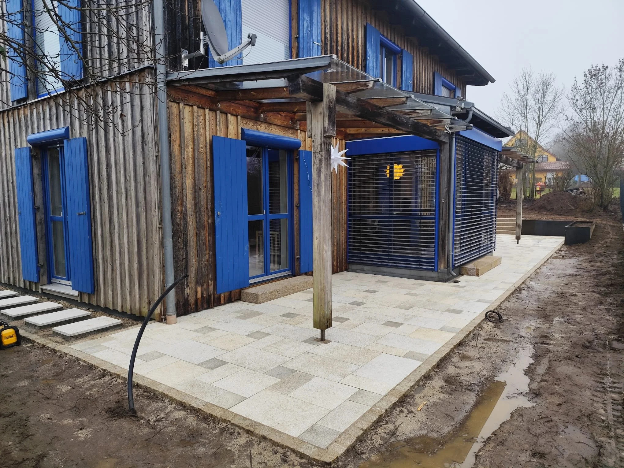 Ansicht eines Hauses mit Holzwand, blauen Fensterläden und einer Terrasse mit gepflastertem Boden, im Hintergrund sind Bäume und ein Nachbarhaus zu sehen.