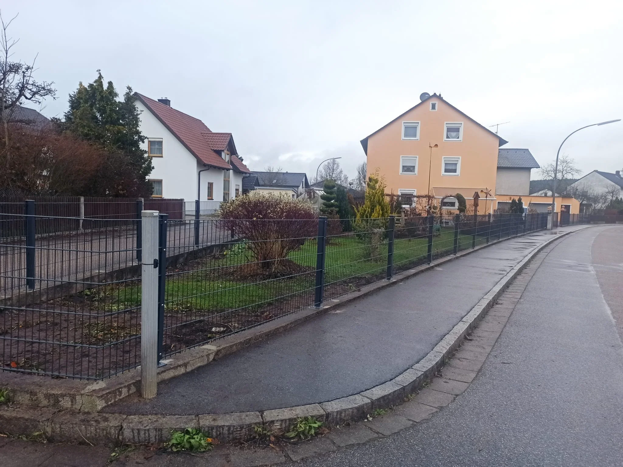 Straße mit Gehweg und Gartenzaun vor mehreren Häusern in einer Wohnsiedlung, bewölkter Himmel, nasse Straße, am Seitenstreifen einige Pflanzen und Sträucher.