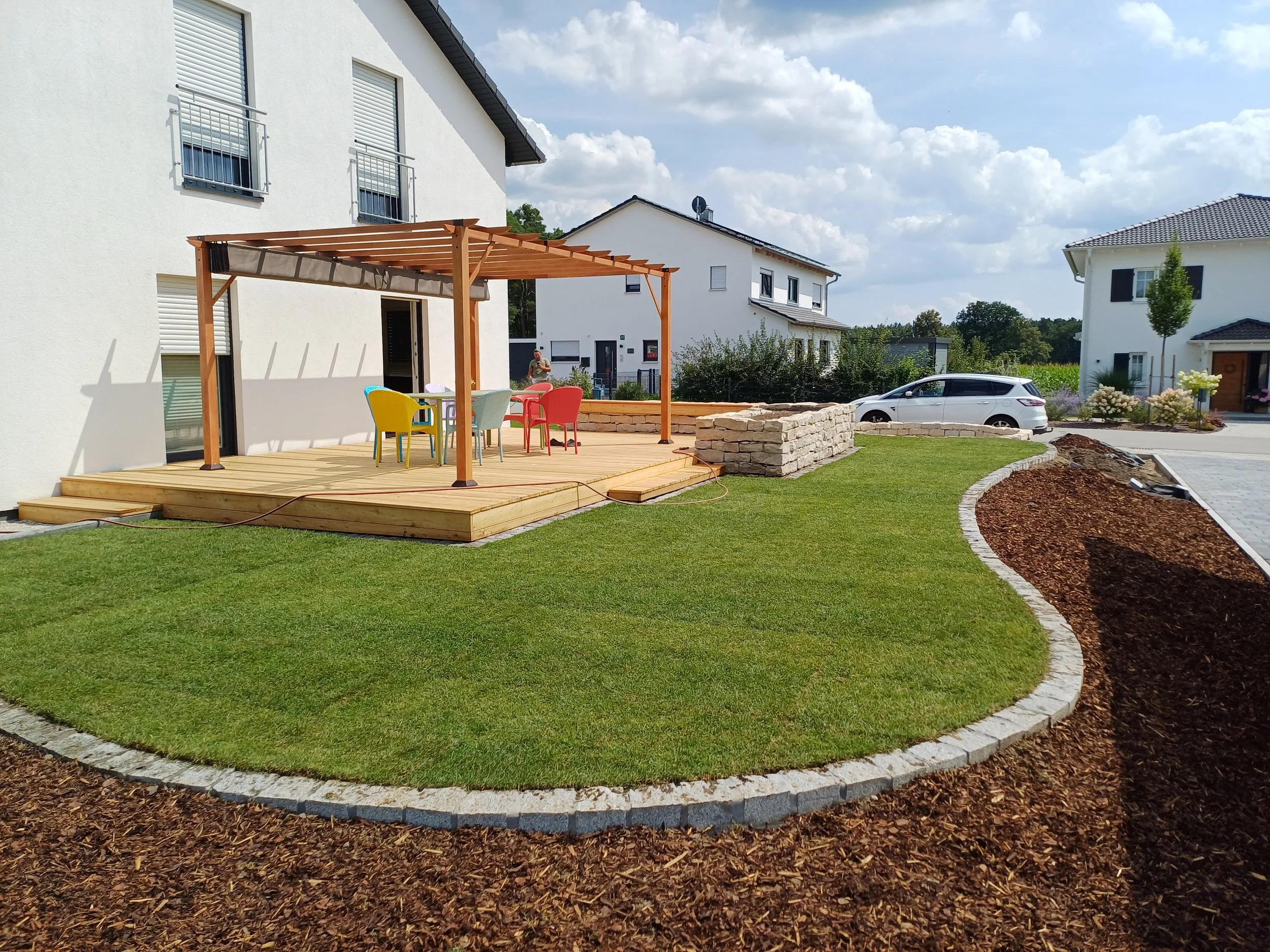 Garten mit Holzterrasse, bunten Stühlen, einem Überdach, grünem Rasen und umrandetem Blumenbeet, mit modernen Häusern im Hintergrund, bei sonnigem Himmel.