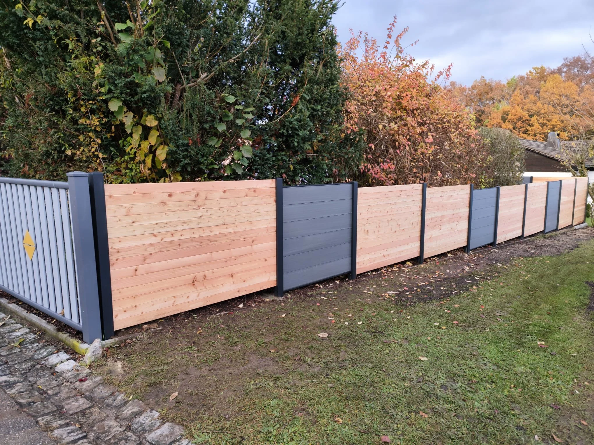 Holzzaun mit blauen Metallpfosten, im Hintergrund Bäume mit Herbstlaub, Rasen und eine kleine Mauer aus Pflastersteinen