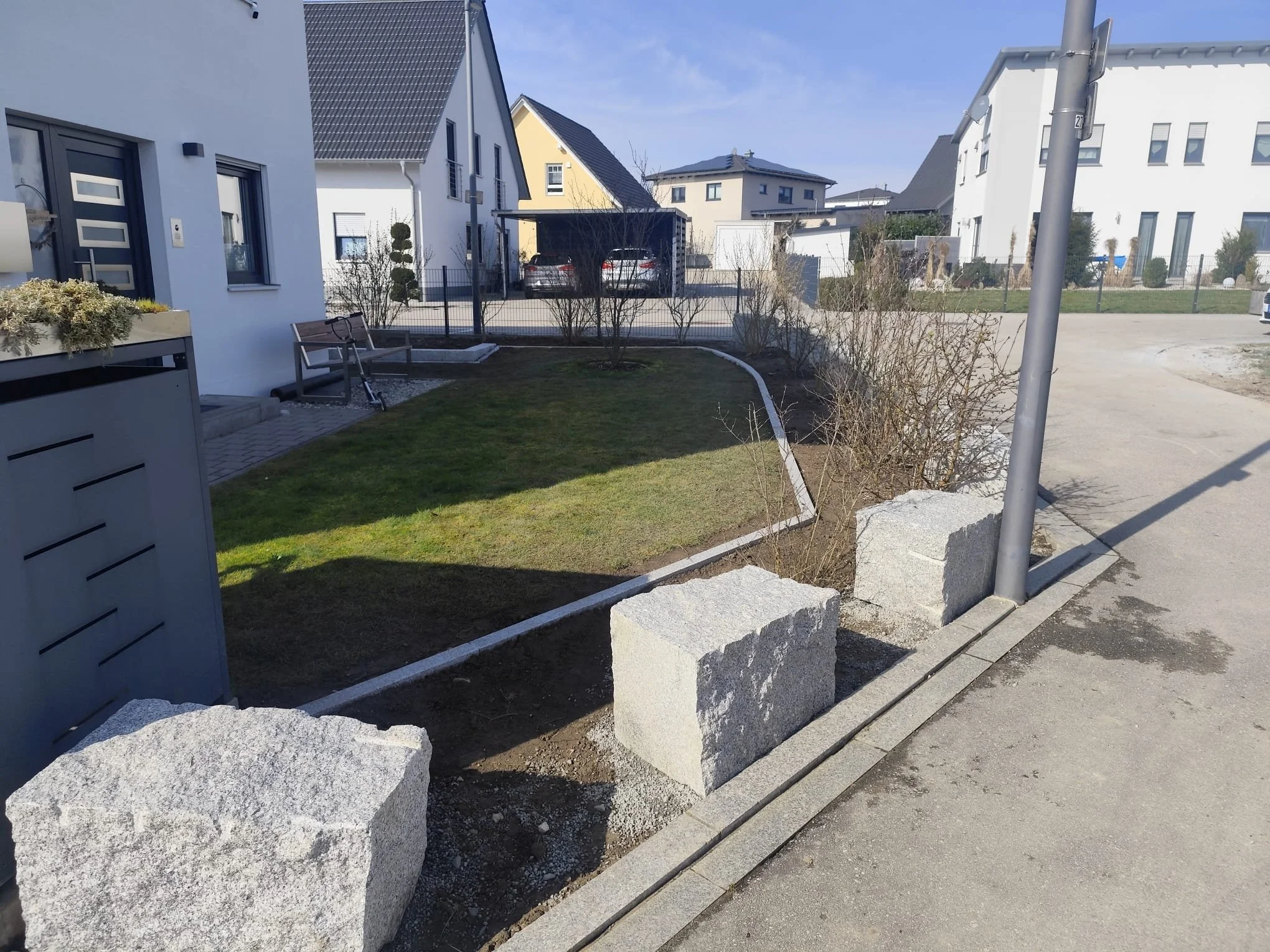 Garten mit Rasen, Sträuchern und einer Bank vor einem weißen Haus. Ein Gehweg und eine Straße mit einem Laternenmast im Vordergrund.
