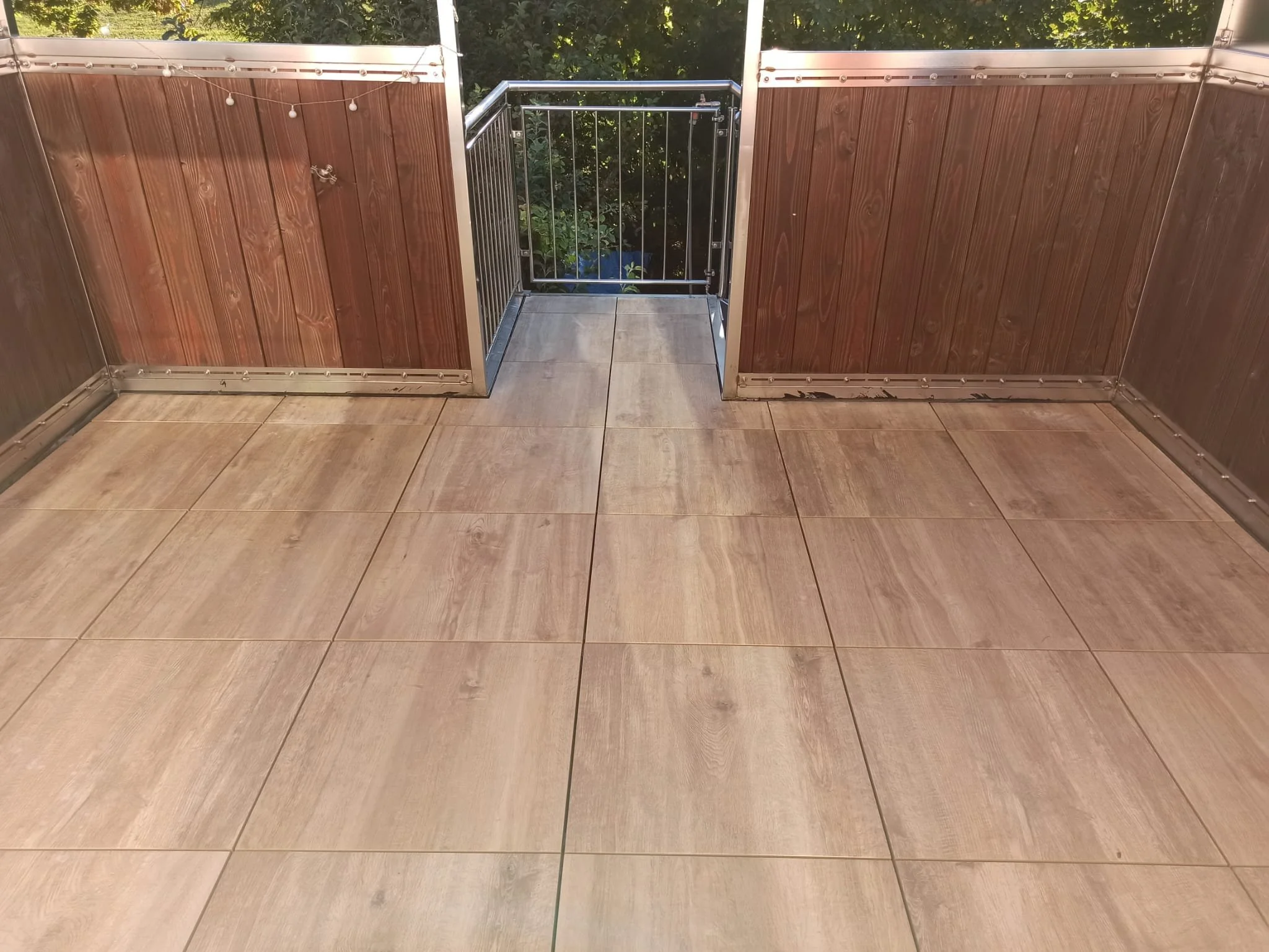 Balkon mit beige Geschossfliesen, Holzverkleidungen und einem kleinen Übergang zur Gartenanlage mit Bäumen im Hintergrund.