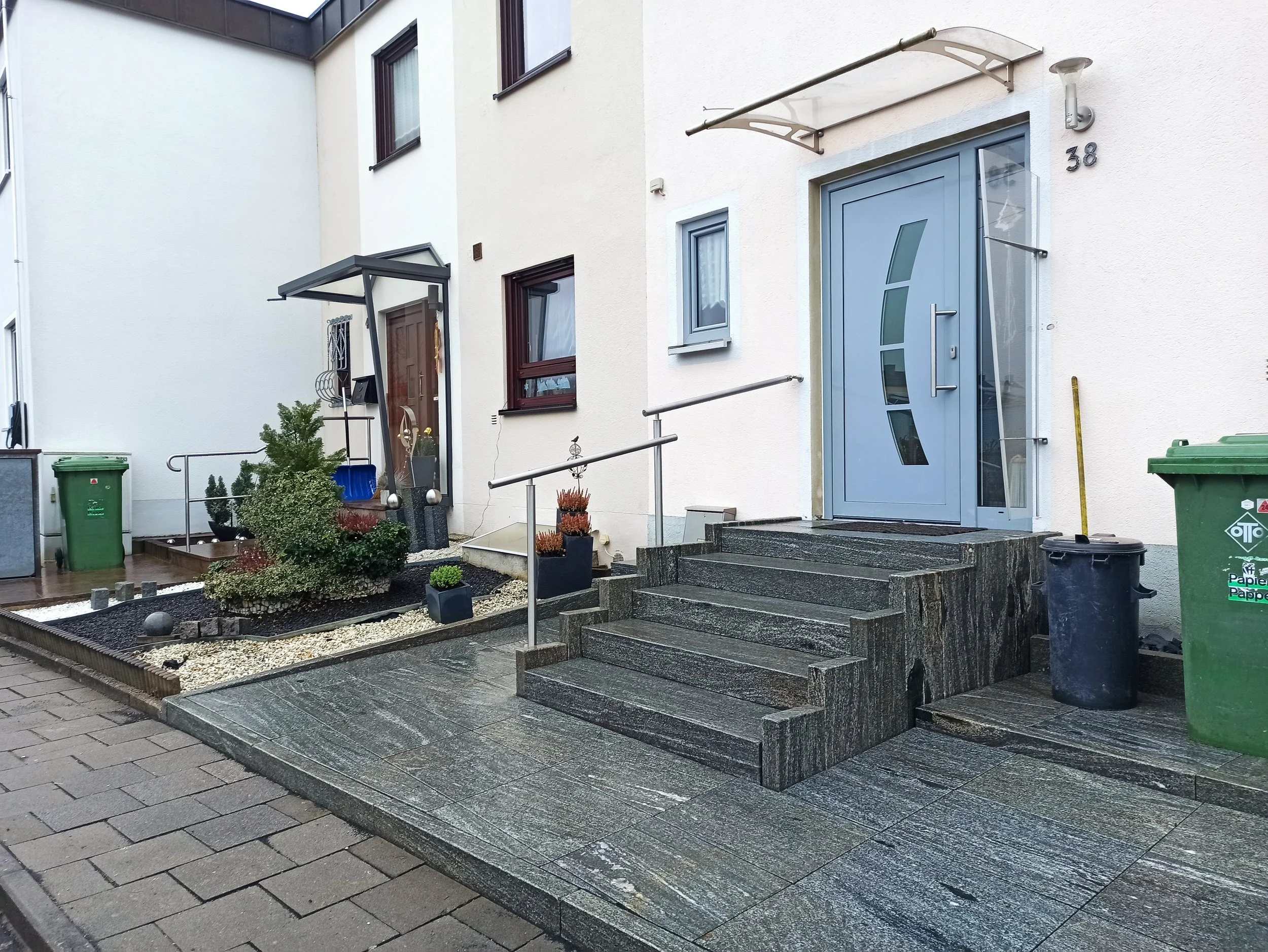 Wohnungseingang mit blauer Tür, Treppen, Garten mit Pflanzen, Mülleimer, und Gehweg in Deutschland.