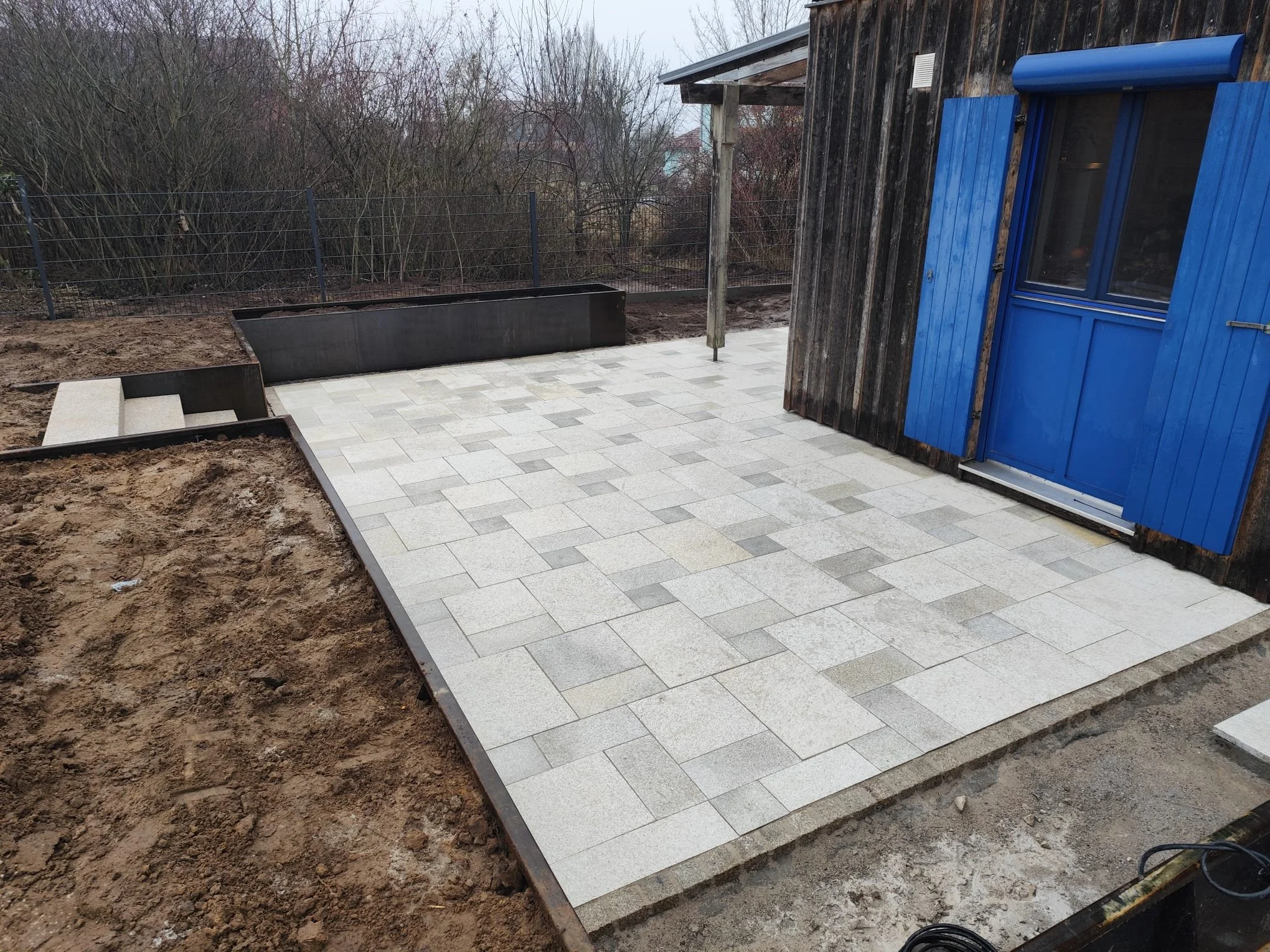 Neue gepflasterte Terrasse neben einem Holzhaus mit blauer Tür und blauen Fensterläden, umgeben von einem Gartenzaun und unberührtem Boden
