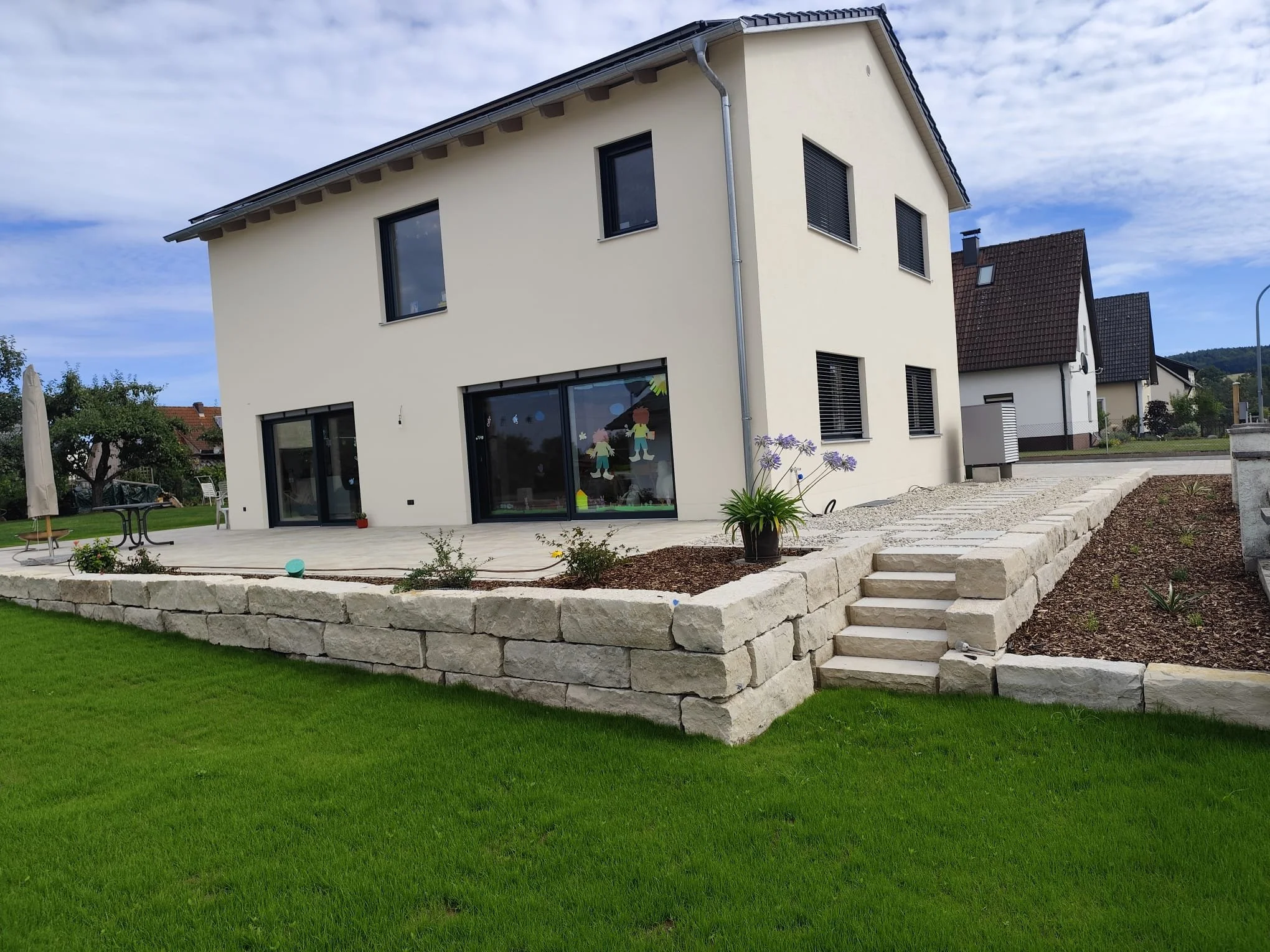 Ein modernes, mehrstöckiges Haus mit weißer Fassade und großen Fenstern, umgeben von einem gepflegten Garten mit grüner Wiese, Steinstufen und einer Terrasse.
