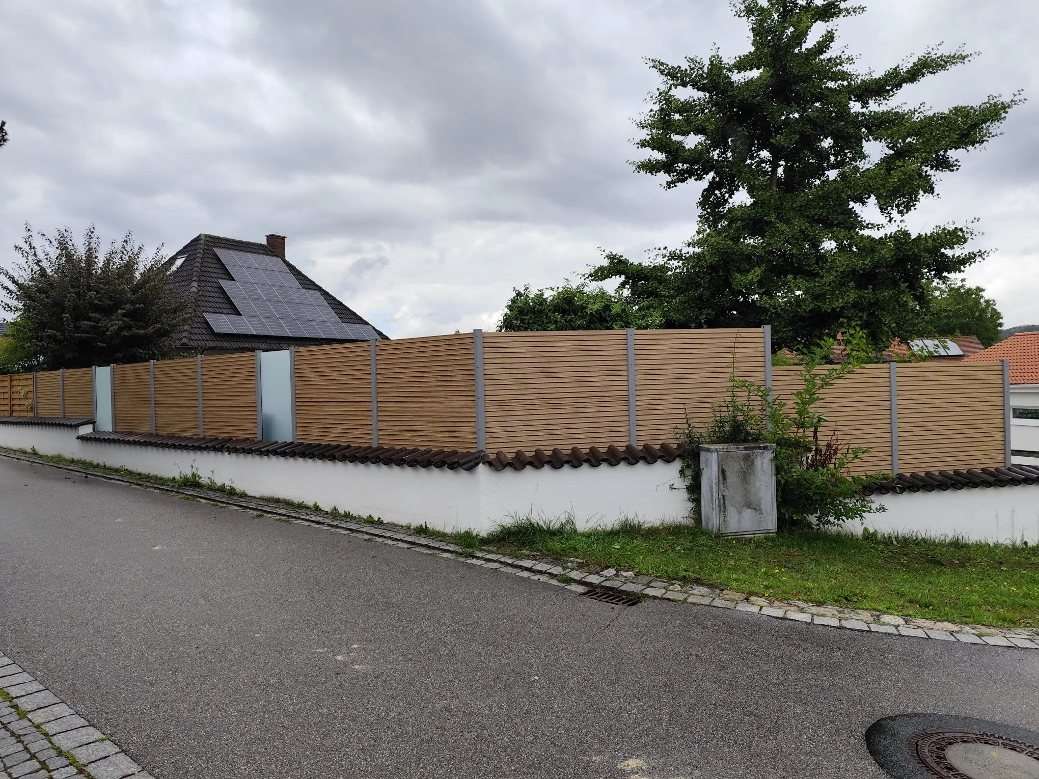 Straße mit einem hohen Holzlattenzaun, Bäumen und einem Haus mit Solarpanelen auf dem Dach im Hintergrund.