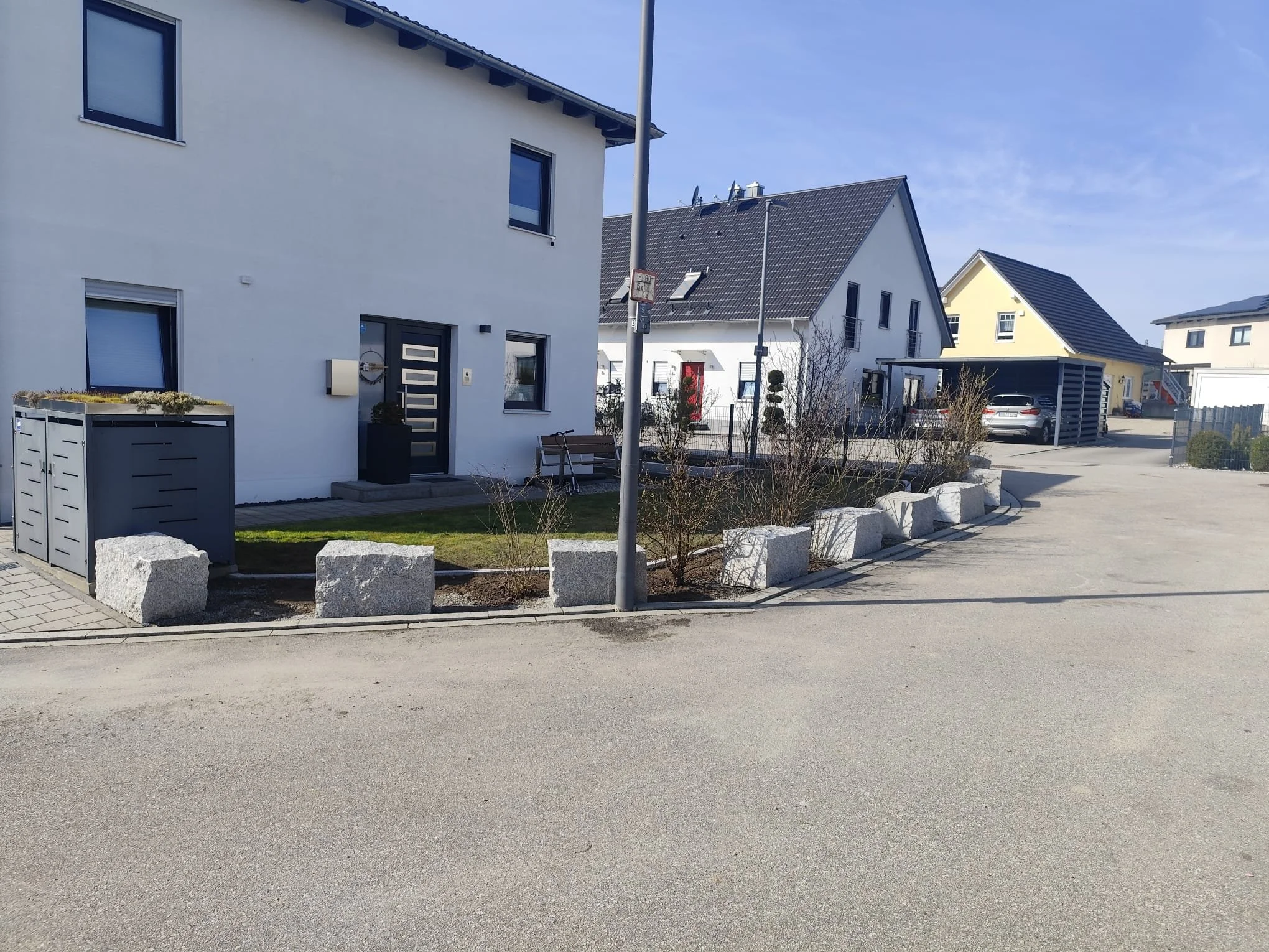 Ein modernes Wohnhaus mit weißer Fassade und schwarzen Fensterrahmen, davor eine kleine Grünfläche mit Steinblöcken und Bänken, im Hintergrund weitere Wohnhäuser und ein blauer Himmel.
