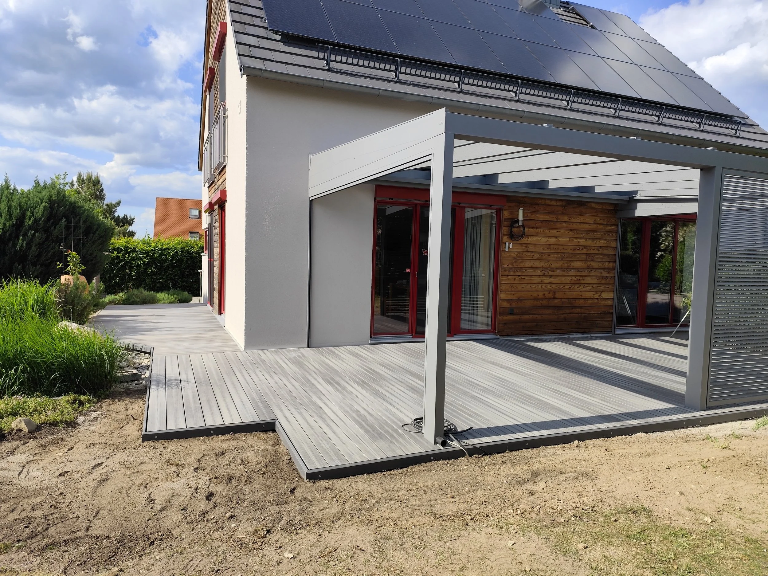Moderne Hausfassade mit Holz- und Glasflächen, Solarzellen auf dem Dach, Holzdeck mit Unterstand im Garten, grüne Bepflanzung im Hintergrund bei sonnigem Himmel.