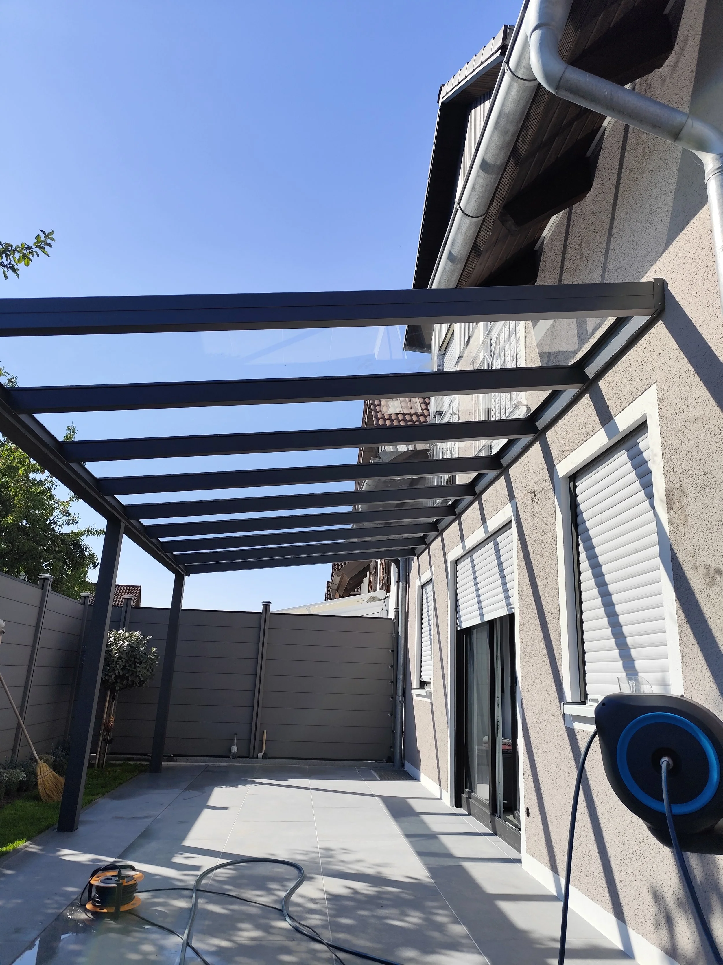Ein Balkon oder Terrasse mit einer modernen Glaskonstruktion als Überdachung, am Tag bei sonnigem Himmel, im Hintergrund Dachbauten und ein blauer Himmel.