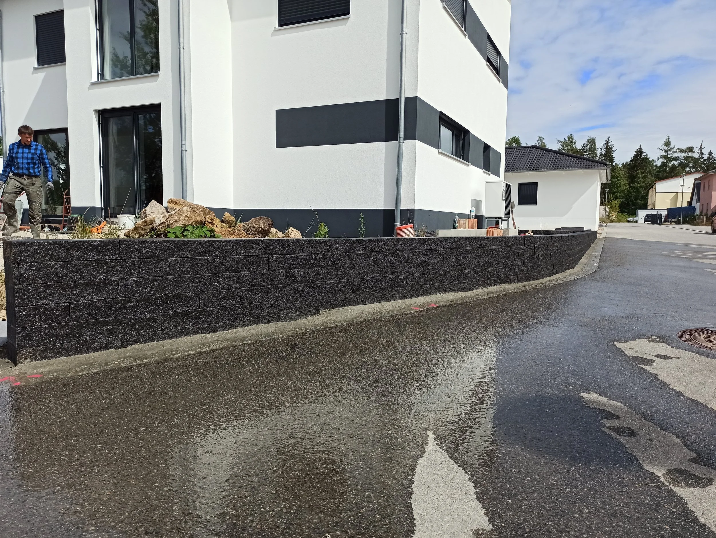 Ein Neubau mit weißer Fassade und schwarzen Akzenten, im Vordergrund ein dunkler Steinmauer, an der Straße. Es sind Bauarbeiten im Gange, mit einem Mann auf der linken Seite, der vor Steinen und Erde steht.