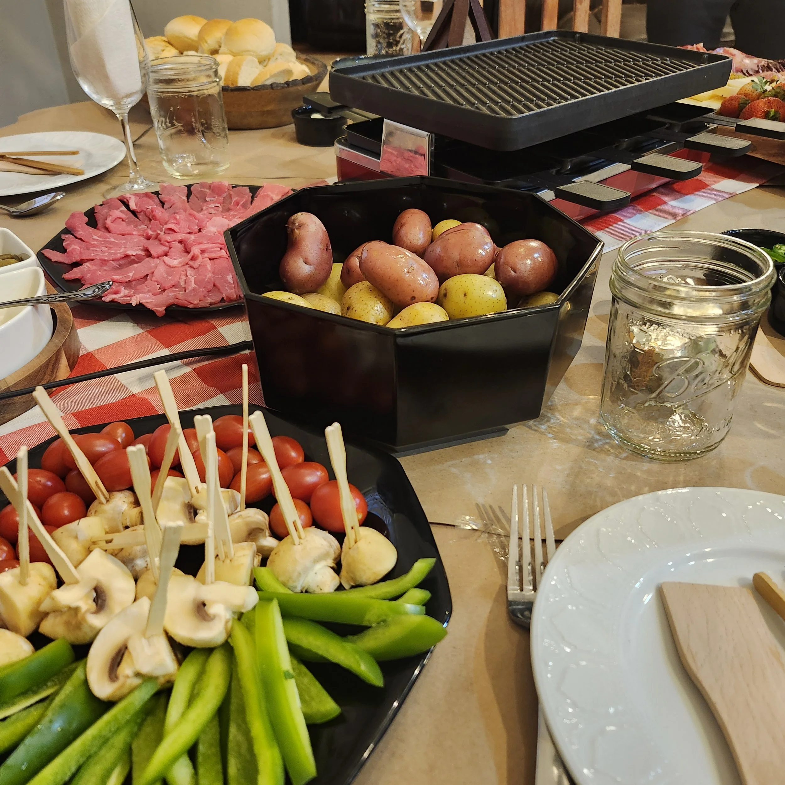 Table set with various foods including raw vegetables, strawberries, bread rolls, sliced meats, and a hot grill for cooking.