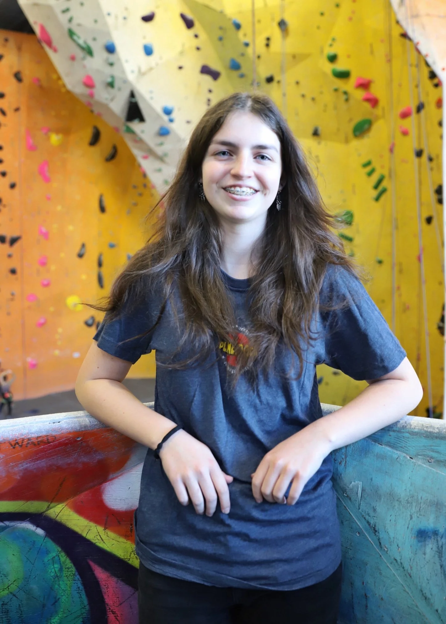A young woman with long brown hair and braces smiling at the camera, standing in front of a colorful indoor rock climbing wall.