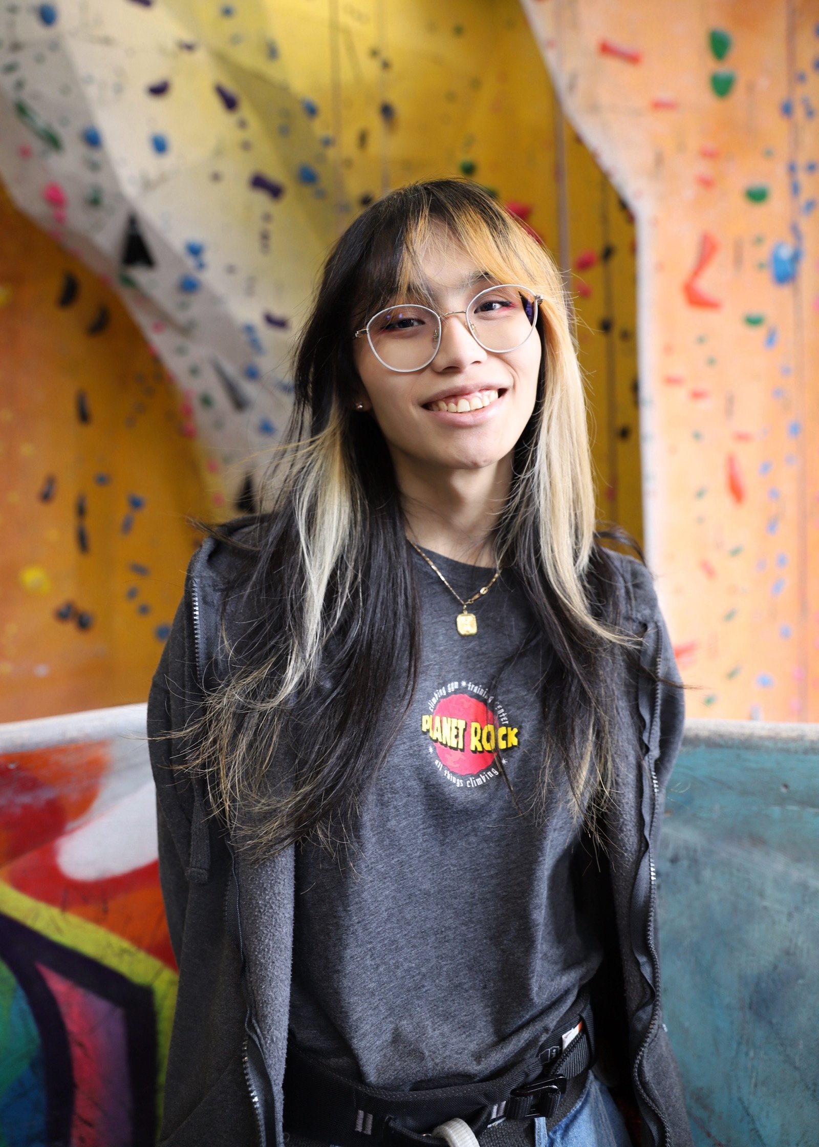 A smiling young person with long black and blonde hair, glasses, wearing a gray "Planet Rock" t-shirt, black jacket, and climbing harness, standing in front of a colorful indoor rock climbing wall.
