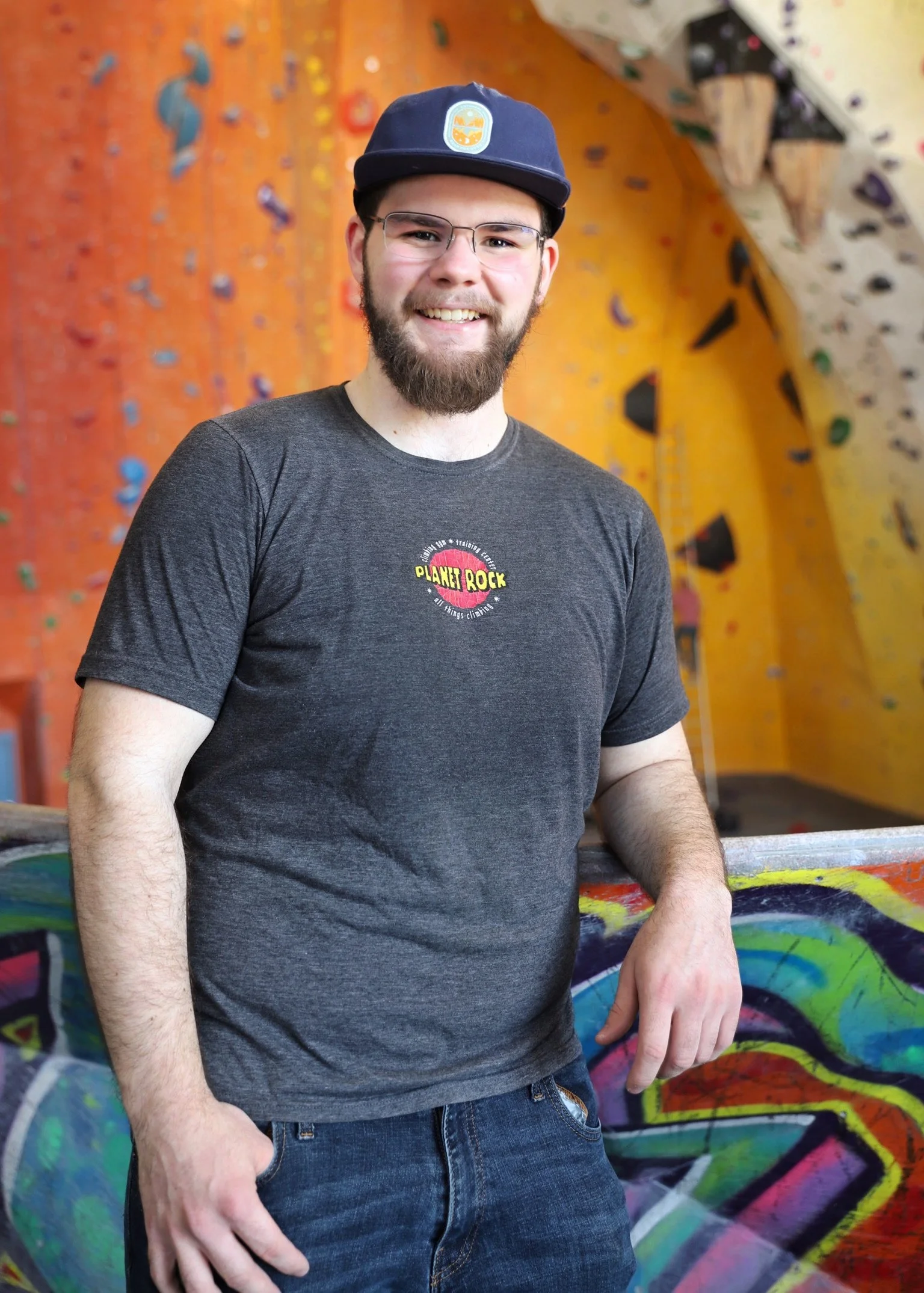 A smiling young man with glasses and a beard, wearing a dark T-shirt with a logo that says "Planet Rock," standing in front of a colorful graffiti wall in an indoor rock climbing gym.