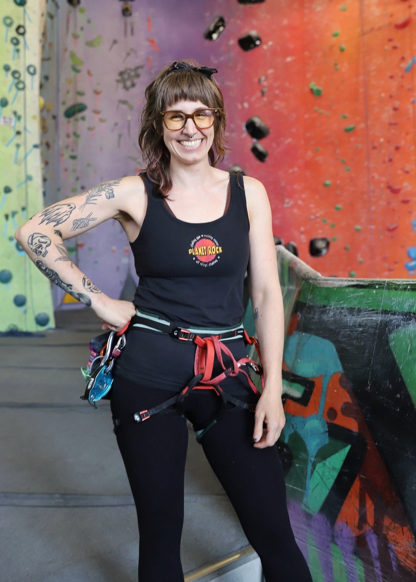 A smiling woman with tattoos on her left arm, wearing glasses and a black tank top with climbing gear around her waist, standing in front of an indoor rock climbing wall.
