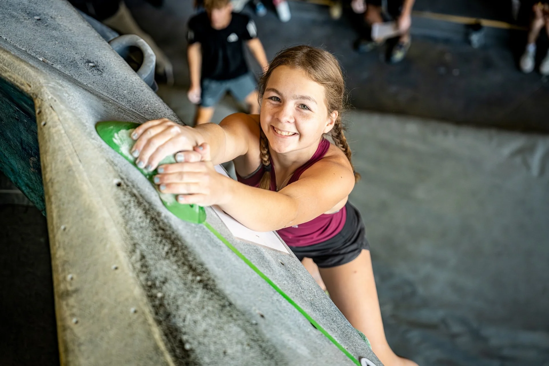 A young girl with braided hair climbing a rock wall at an indoor climbing gym, smiling at the camera.