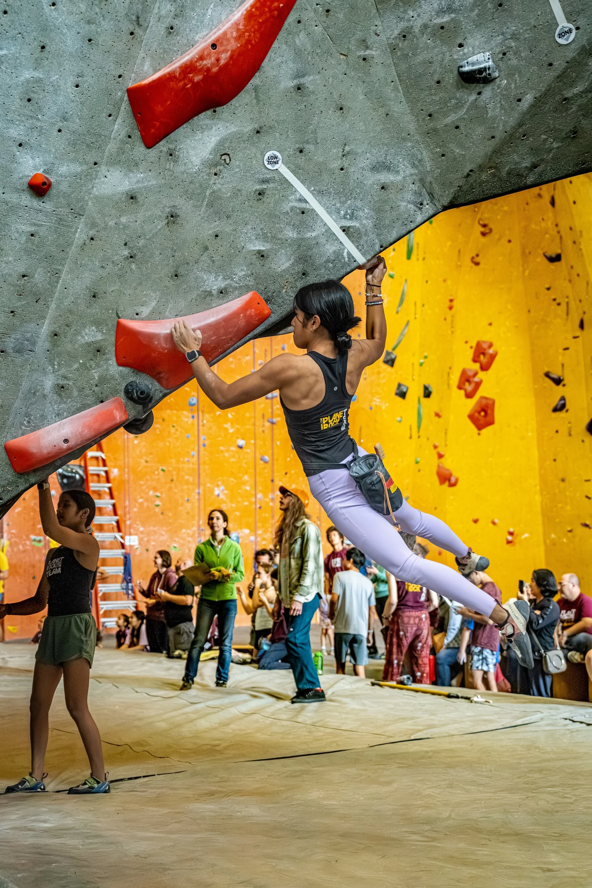 An indoor rock climbing gym with many climbers and spectators. A woman is bouldering, reaching for a hold on an overhanging wall. She is wearing white pants, a black tank top, and climbing shoes. People are observing and engaging with the climbing activity.