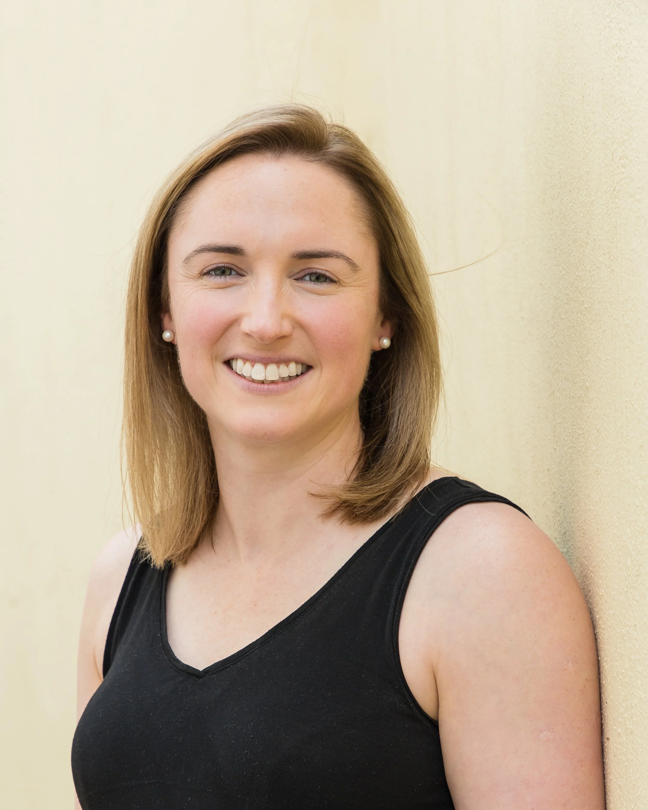 A smiling woman with shoulder-length light brown hair, wearing pearl earrings and a black sleeveless top, standing against a light yellow wall.