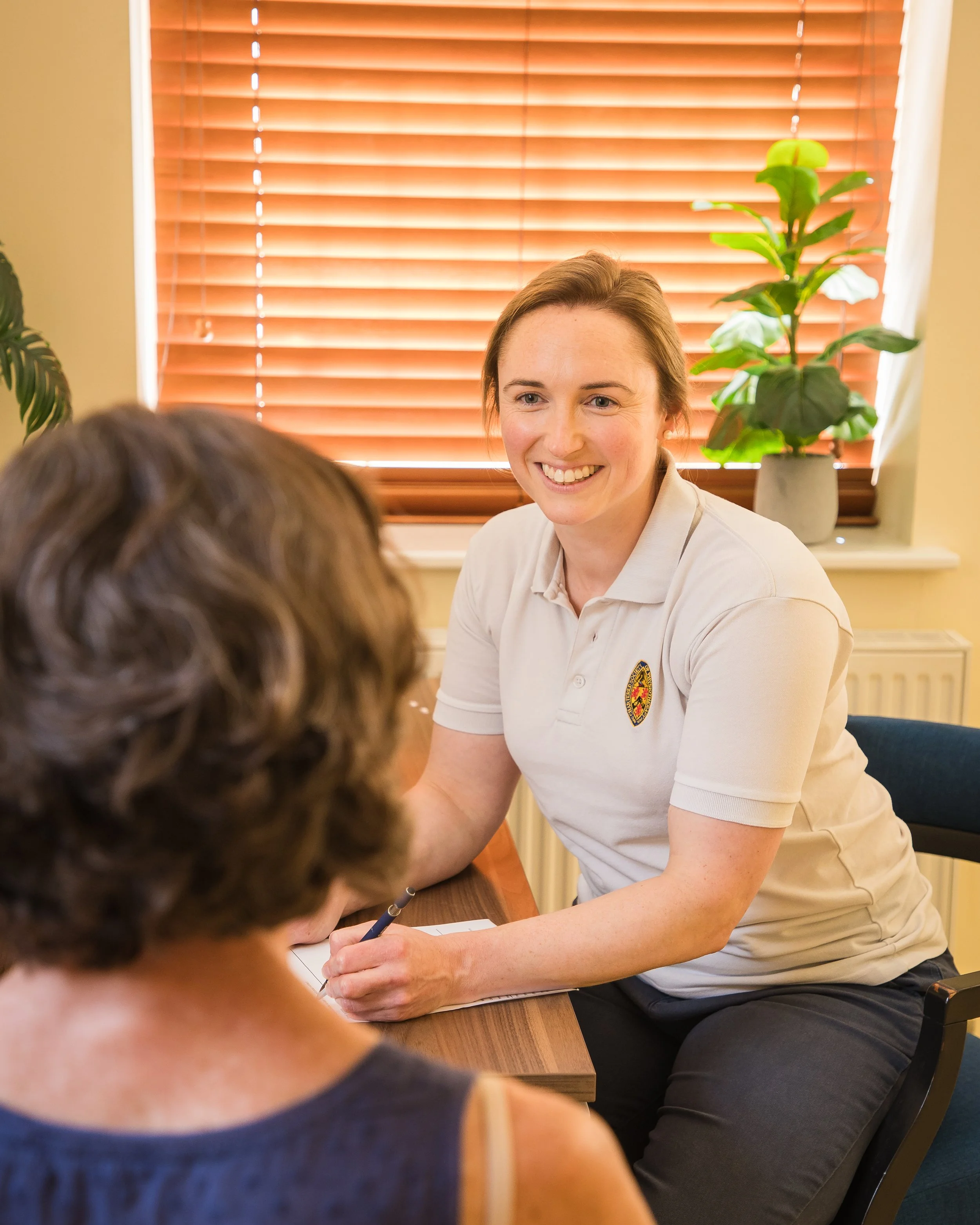 A woman wearing a white polo shirt with an emblem on it, smiling and talking to a person with curly hair. They are sitting at a wooden table in a room with wooden blinds and potted plants.