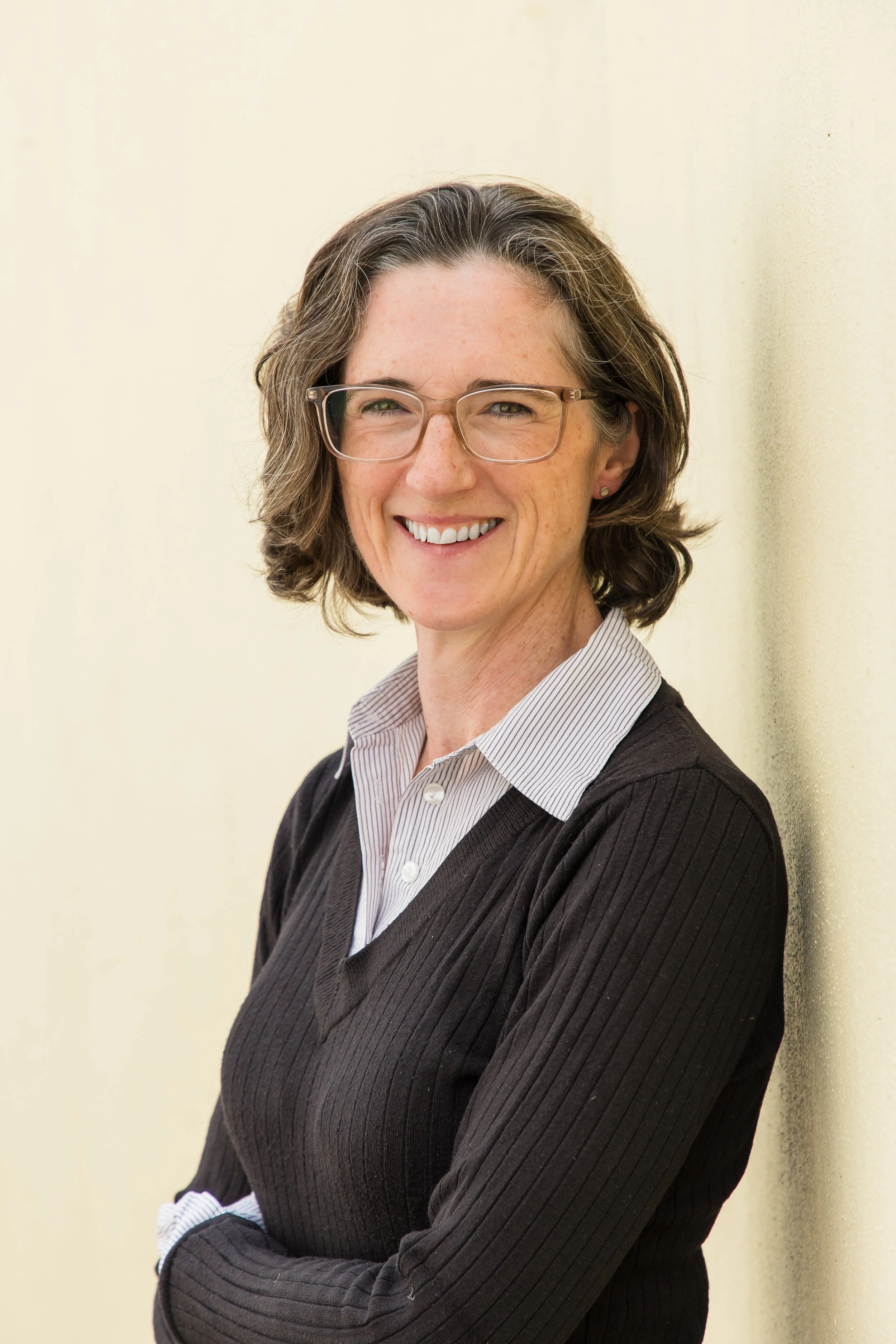 A woman with shoulder-length brown hair, wearing glasses, a striped collared shirt, and a dark sweater, smiling and leaning against a light-colored wall.