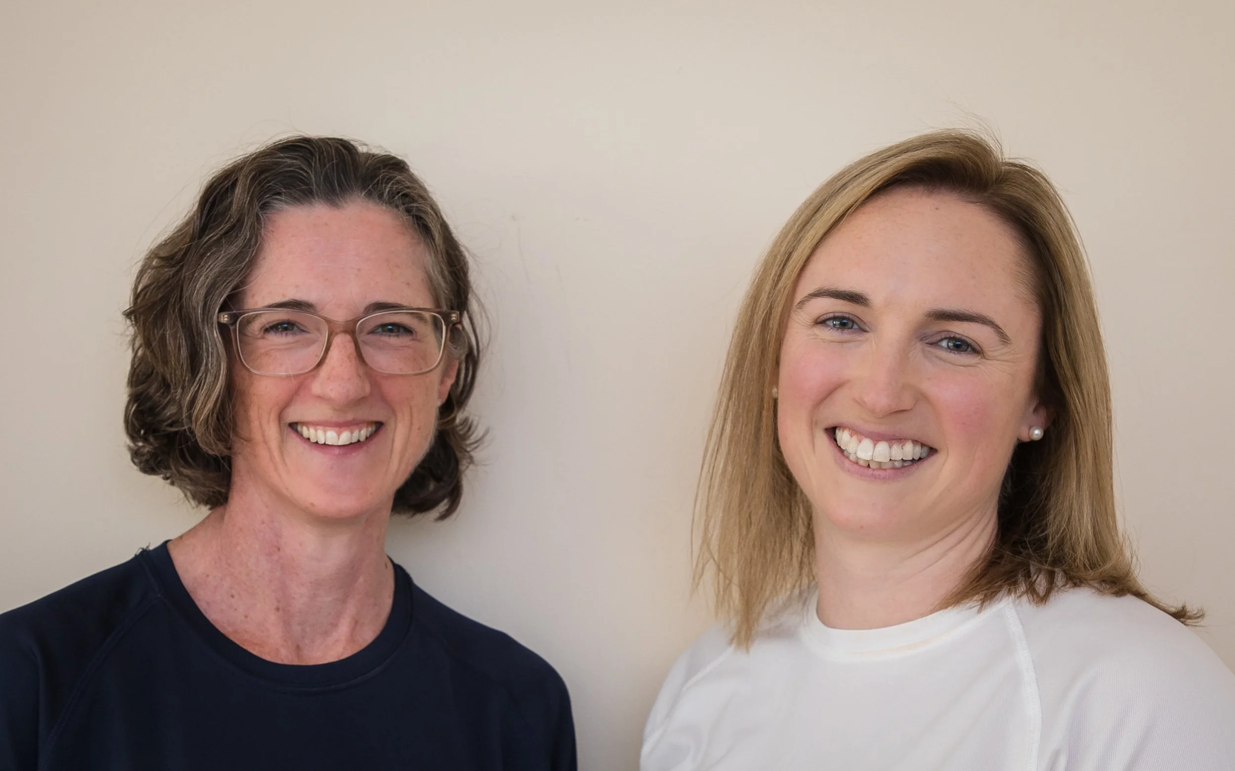 Two women smiling and standing close together in front of a plain light-colored wall, one with glasses and curly hair, the other with straight hair and pearl earrings.