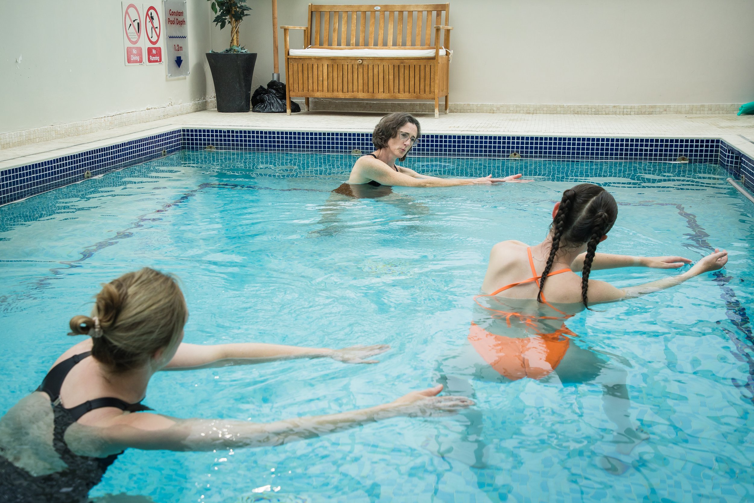 A woman in a black swimsuit and glasses is instructing a young girl with braided hair and an orange swimsuit, both practicing water exercise in a swimming pool. There are signs on the wall indicating pool rules and a wooden bench on the pool deck.