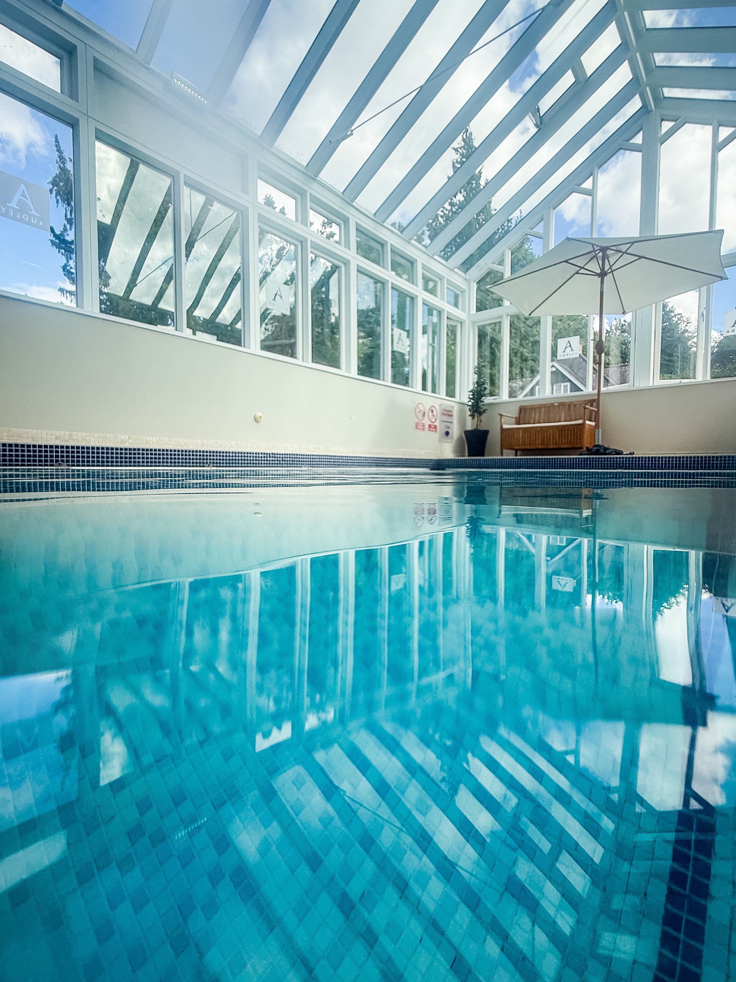 Indoor swimming pool with blue tiles, glass ceiling, and large windows showing trees outside; pool reflections visible.