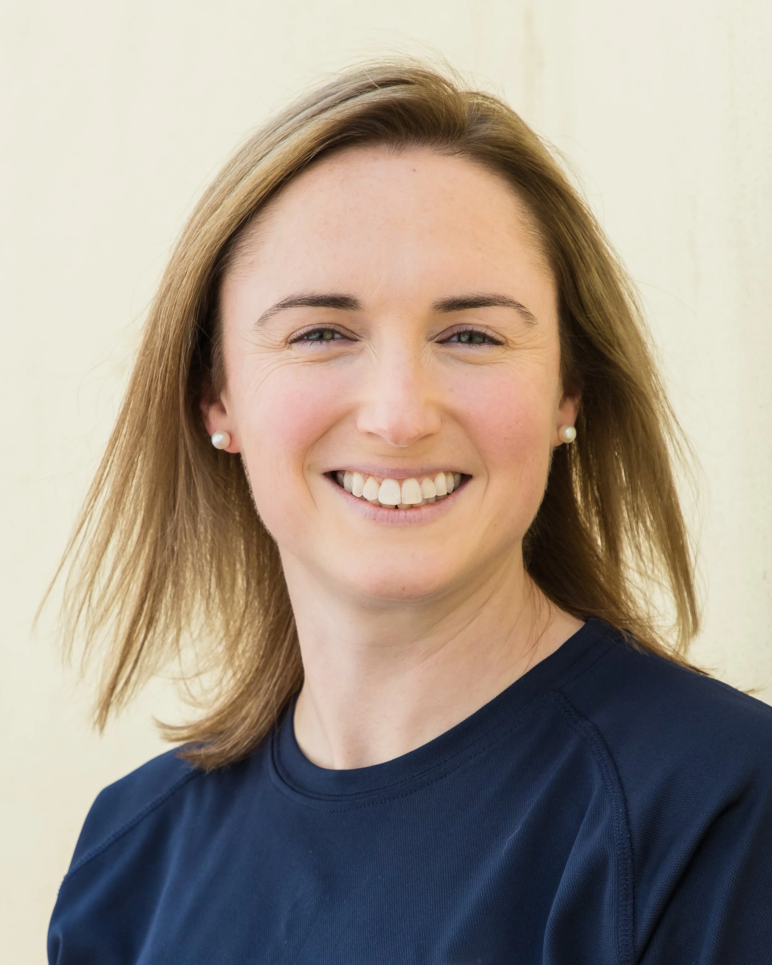 Close-up of a young woman with long, light brown hair smiling, wearing pearl earrings and a navy blue top, standing in front of a light-colored wall.