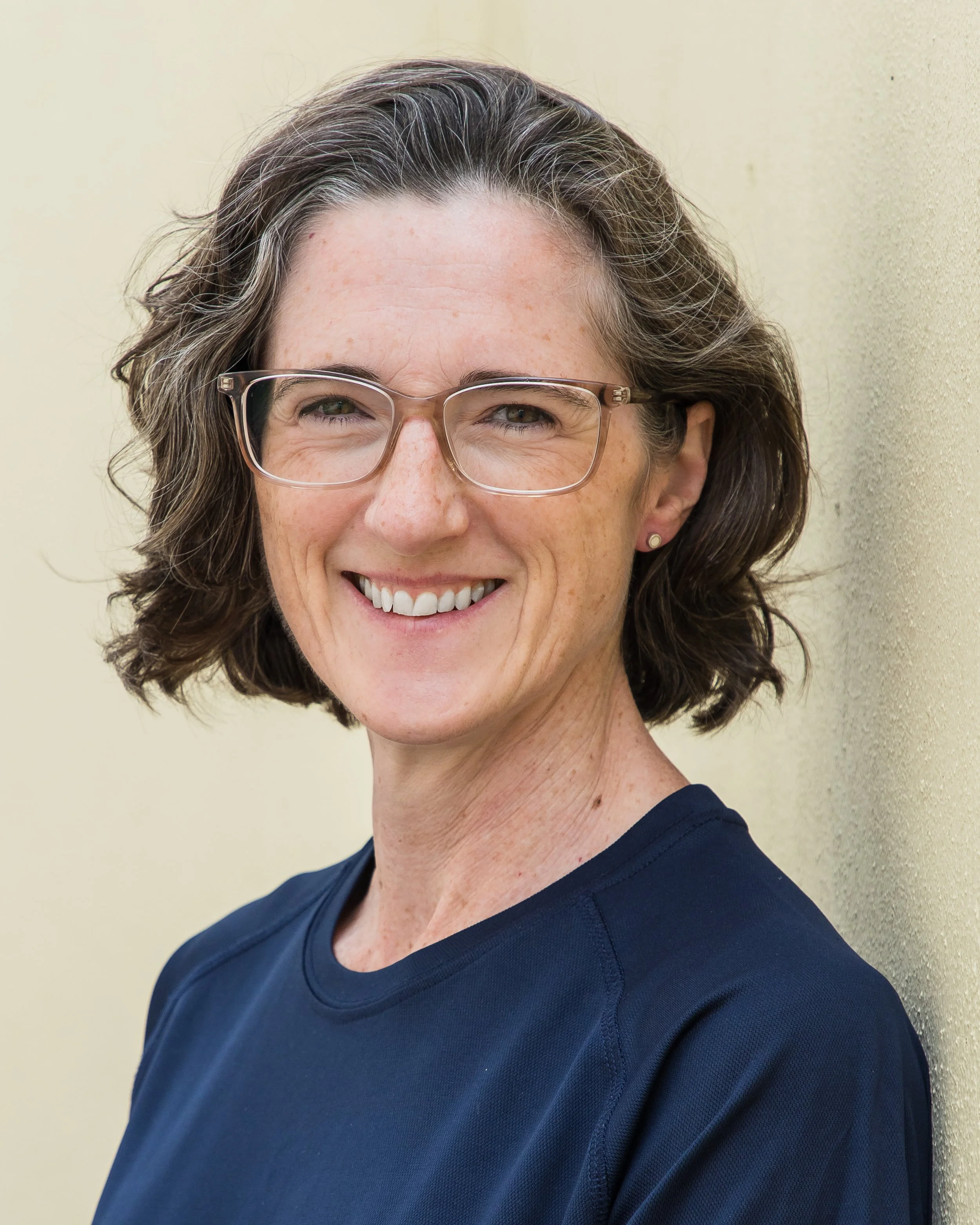 Close-up of a woman with short, wavy brown hair wearing glasses and a dark blue top, smiling against a light beige wall.