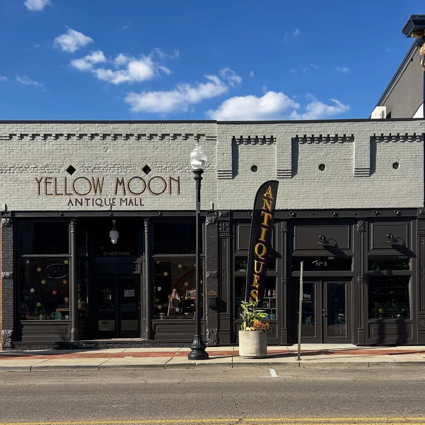 Exterior of Yellow Moon Antique Mall with black storefront, large windows, sign, and a vertical flag banner. Bright daytime with a blue sky and few clouds.