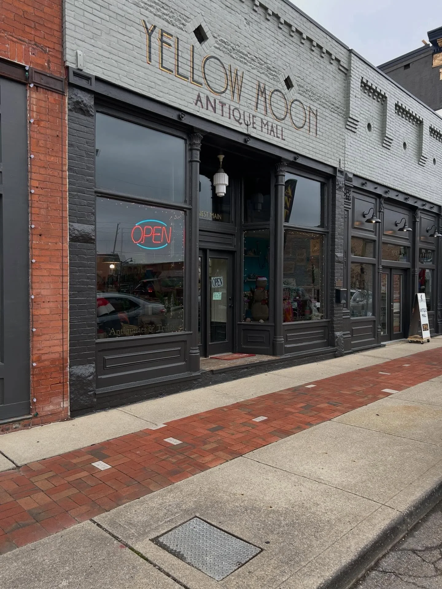 The storefront of Yelow Moon Antique Mall with a brick and black painted exterior, large windows, an illuminated 'OPEN' sign, and a sidewalk in front.