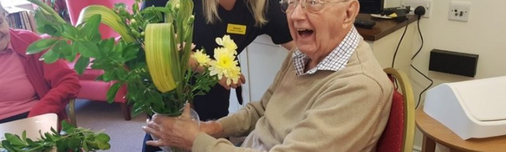 An elderly gentleman sitting in a chair, holding flowers, and smiling