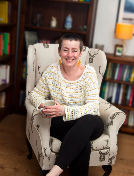 Leesa is sitting in a wingback chair, holding a mug, legs crossed, smiling straight into the camera. There are bookshelves behind her