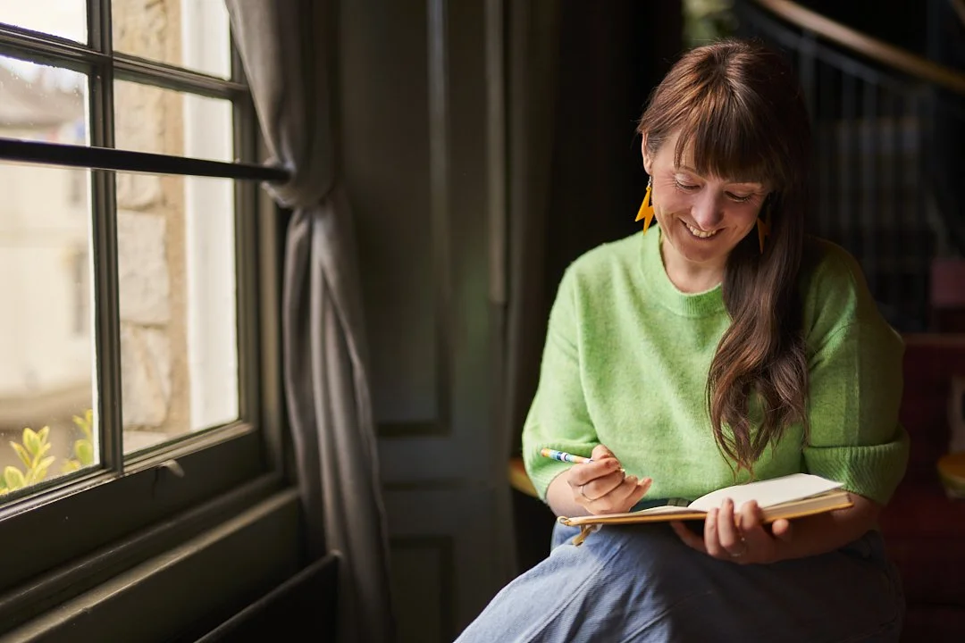 Leesa is wearing a green jumper and blue jeans. She is sitting by a window writing in a notebook.