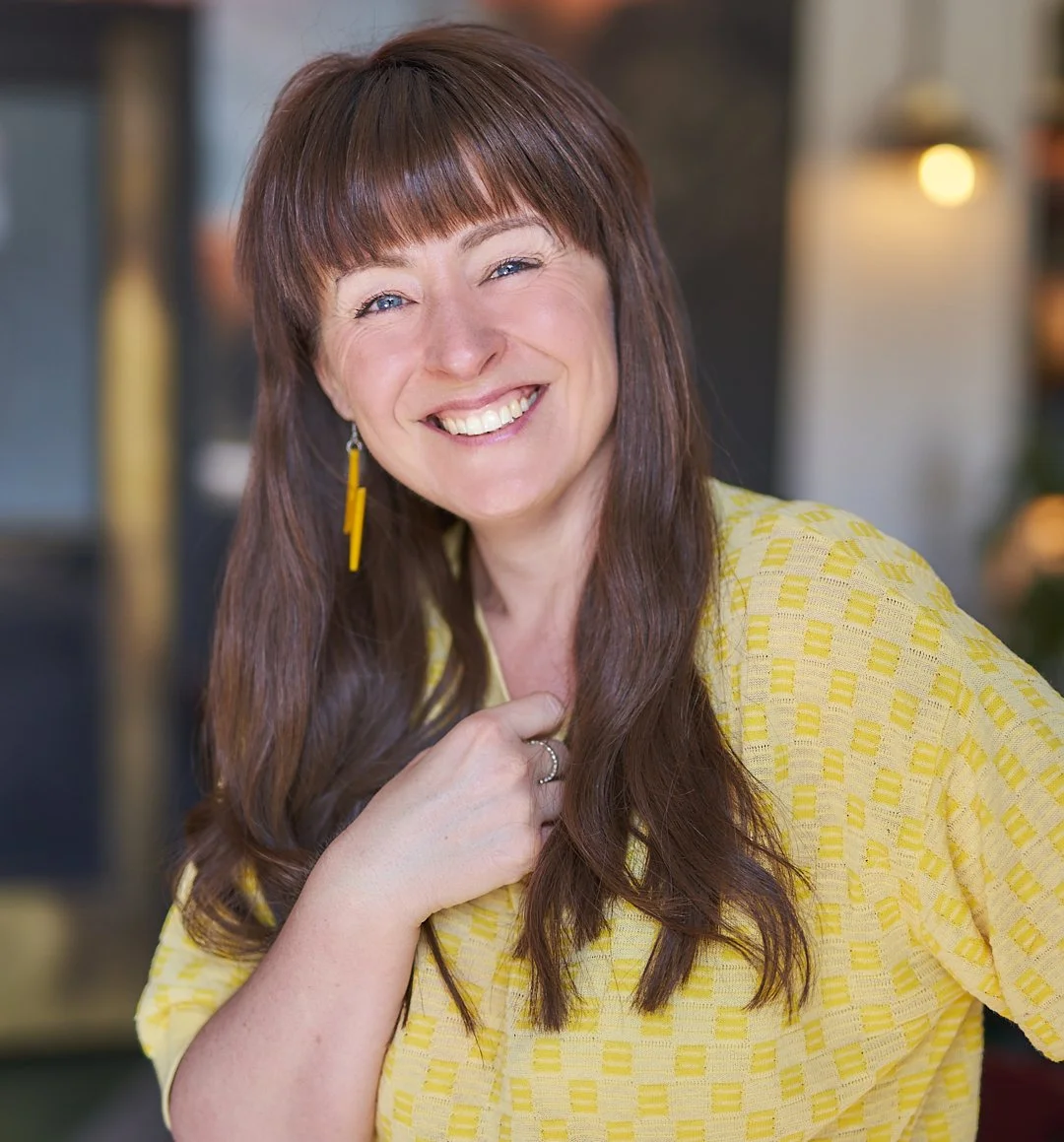 Leesa is standing and smiling at the camera, she has long brown hair and is wearing a yellow t-shirt.