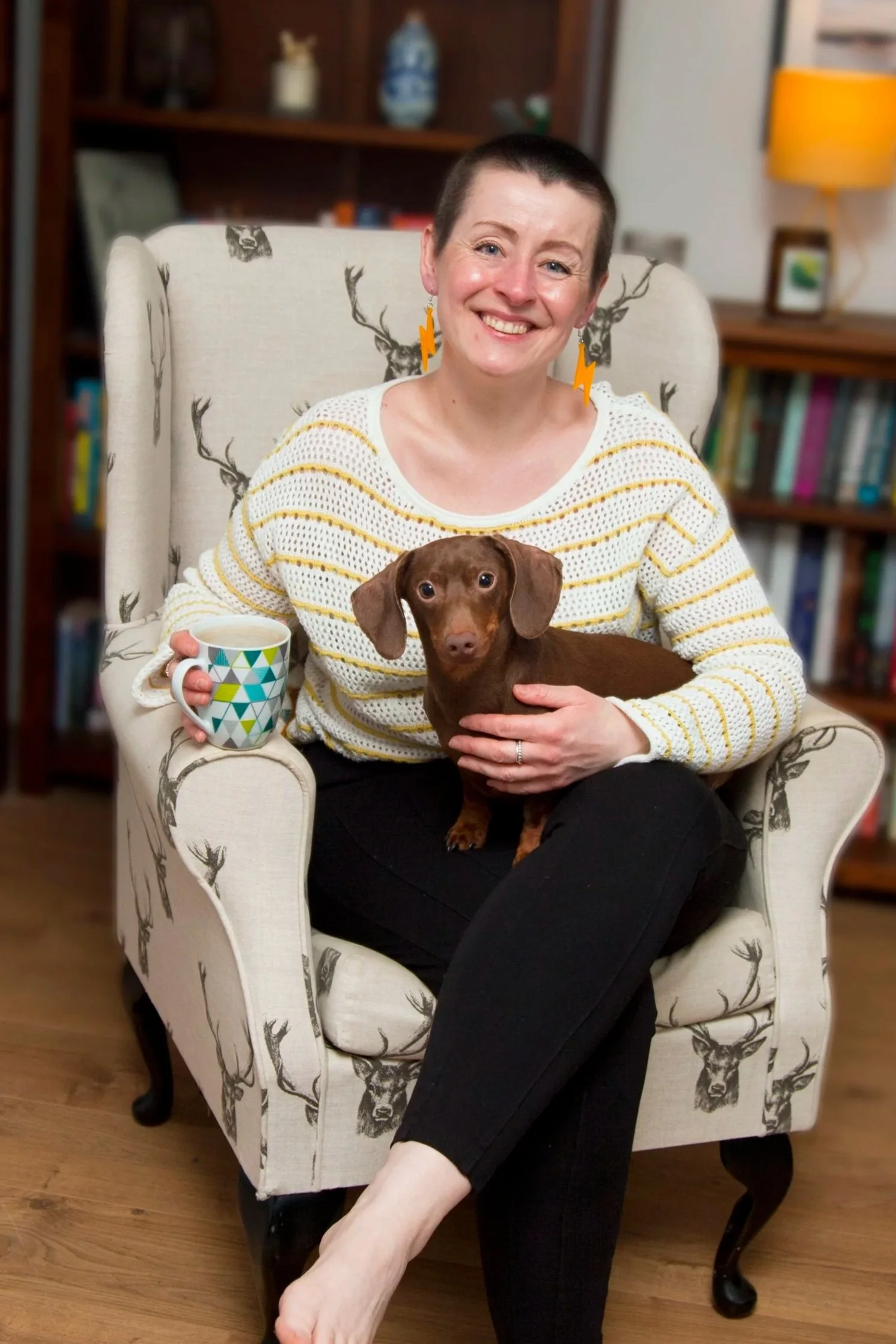 A woman with short hair and earrings shaped like lightning bolts sitting in a beige armchair with deer print, holding a small brown dachshund on her lap and a colorful mug in her right hand, inside a room with bookshelves and a lamp in the background.