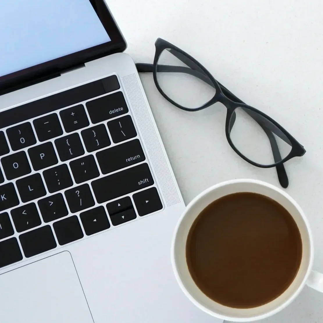 Laptop keyboard, eyeglasses, cup of coffee on a white surface.