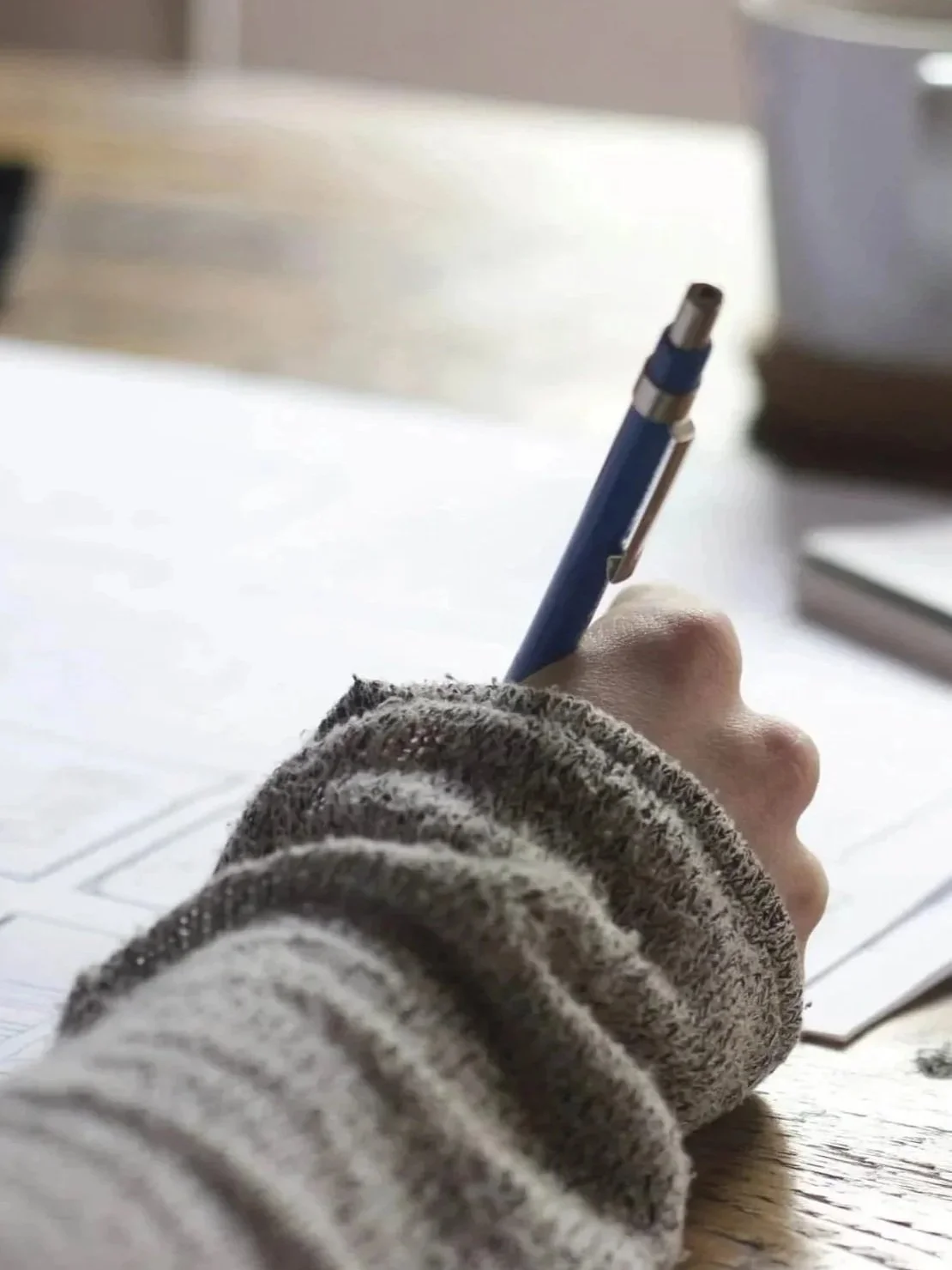 A person's hand holding a blue pen, resting on a desk with papers.