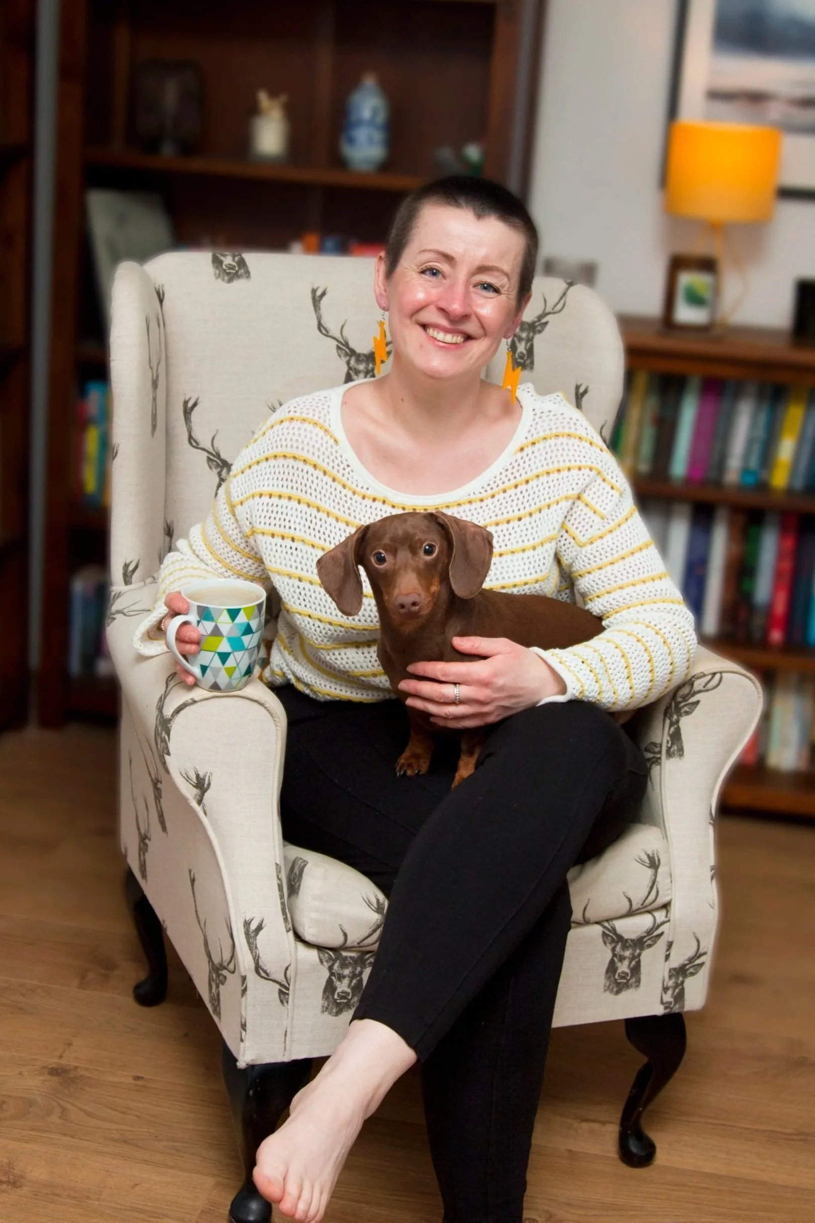 A woman with short hair sitting in a wingback armchair with a dachshund on her lap, holding a coffee mug, in a cozy living room.