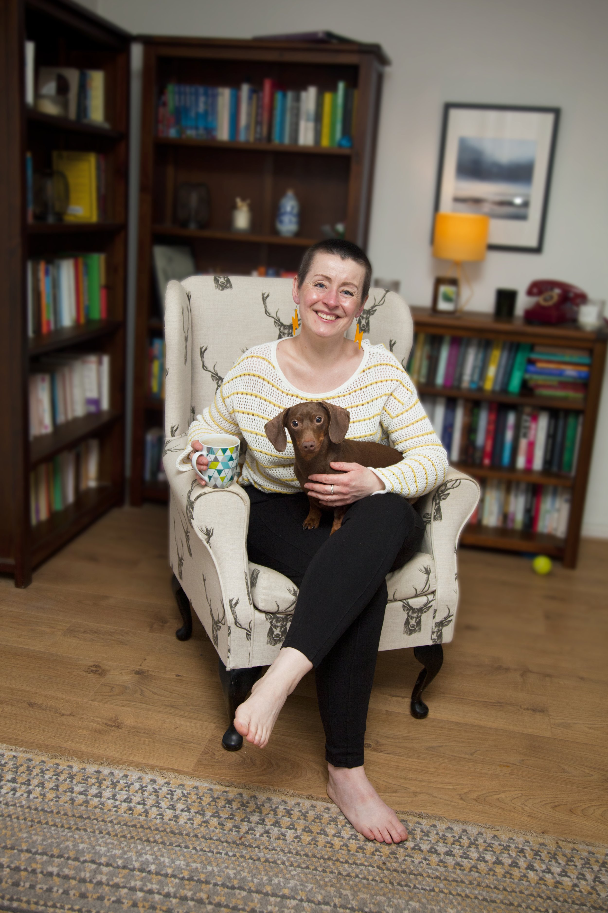 Leesa is sitting in a cream wingback chair, holding a mug of tea. Pluto, the miniature chocolate dachshund is sitting on her lap. There are bookshelves full of books in the background.